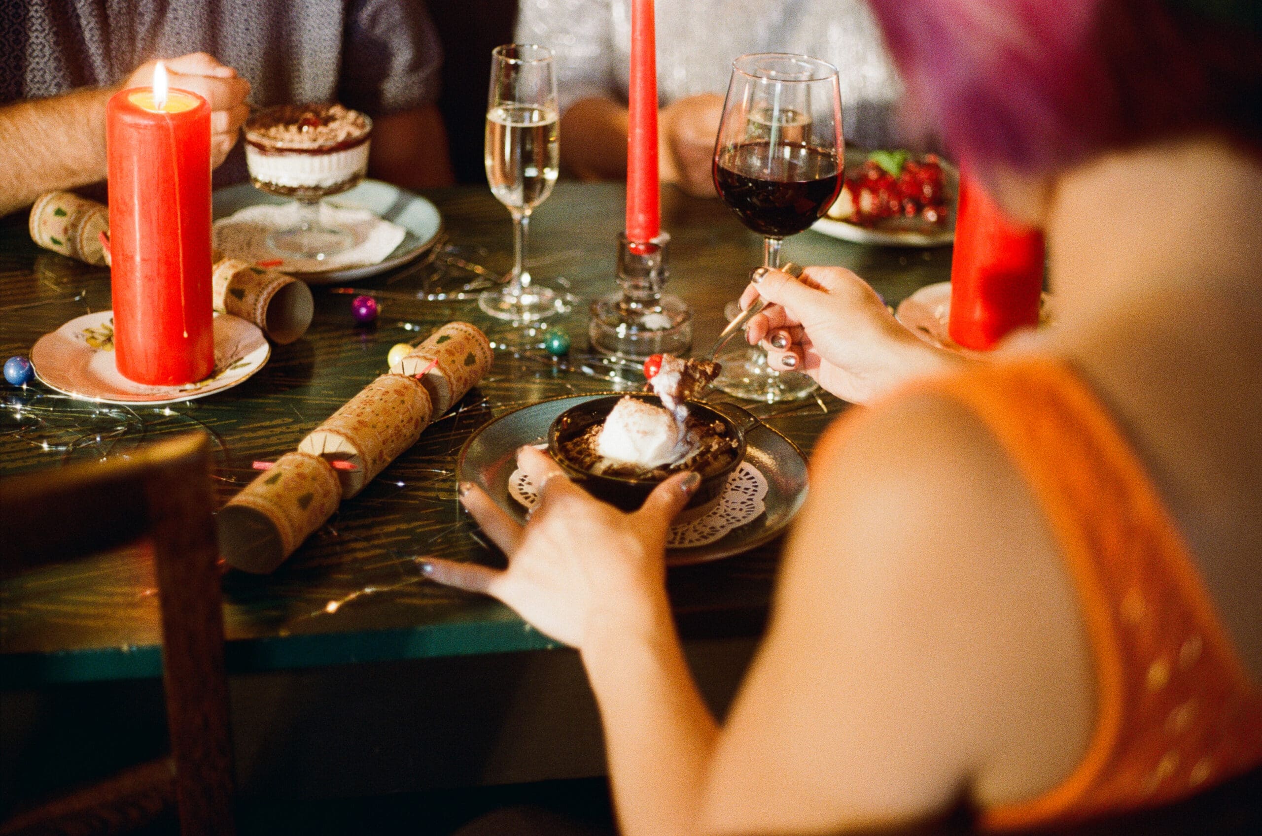 A person in an orange top holds a dessert topped with whipped cream and a cherry at a festive dinner table with red candles, drinks, and holiday decorations. Other people and treats are also visible.