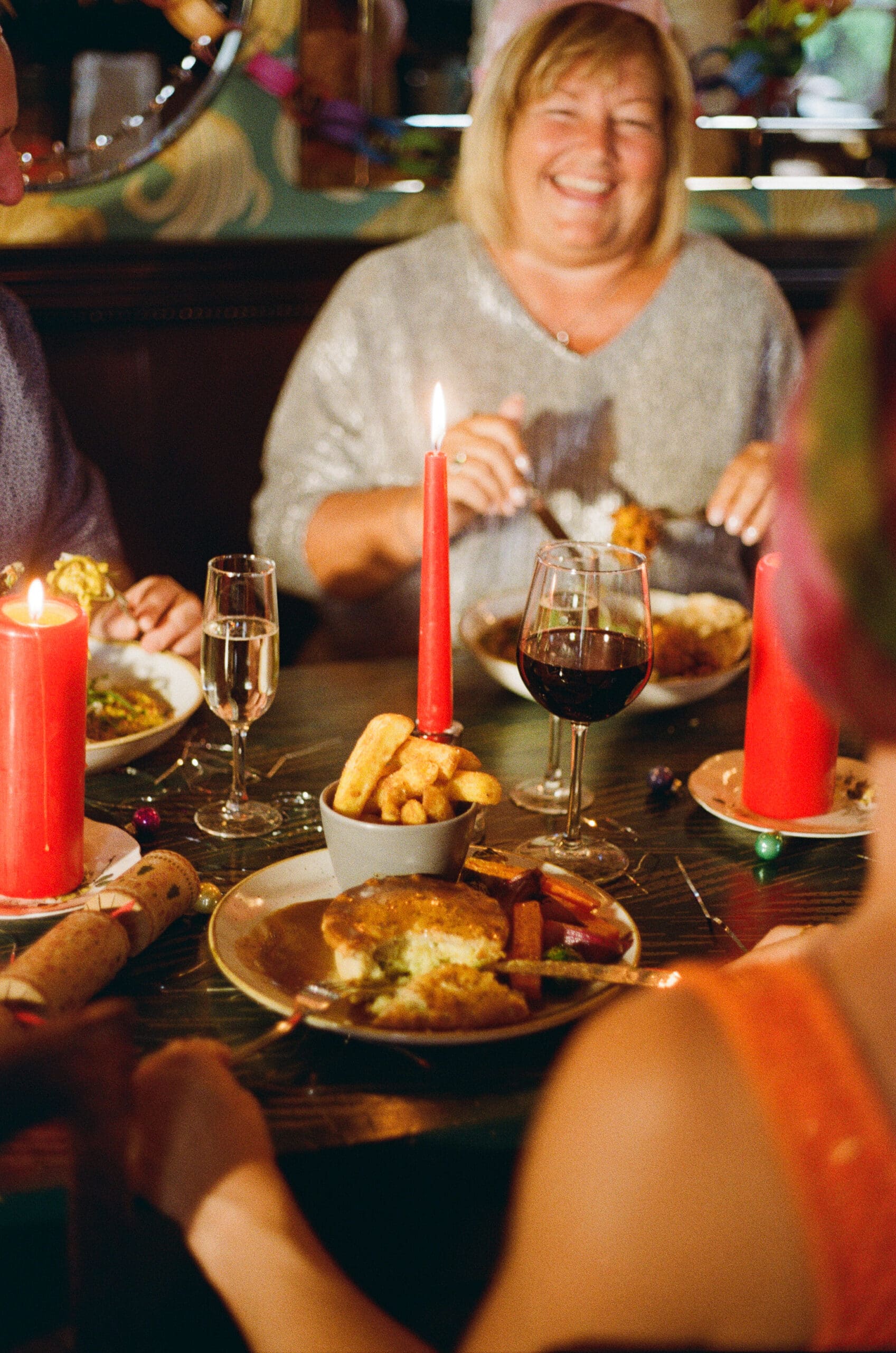 A woman smiling at a festive dinner table set with red candles, drinks, and plates of food. Other people are partially visible, enjoying the meal in a warm, joyful atmosphere.