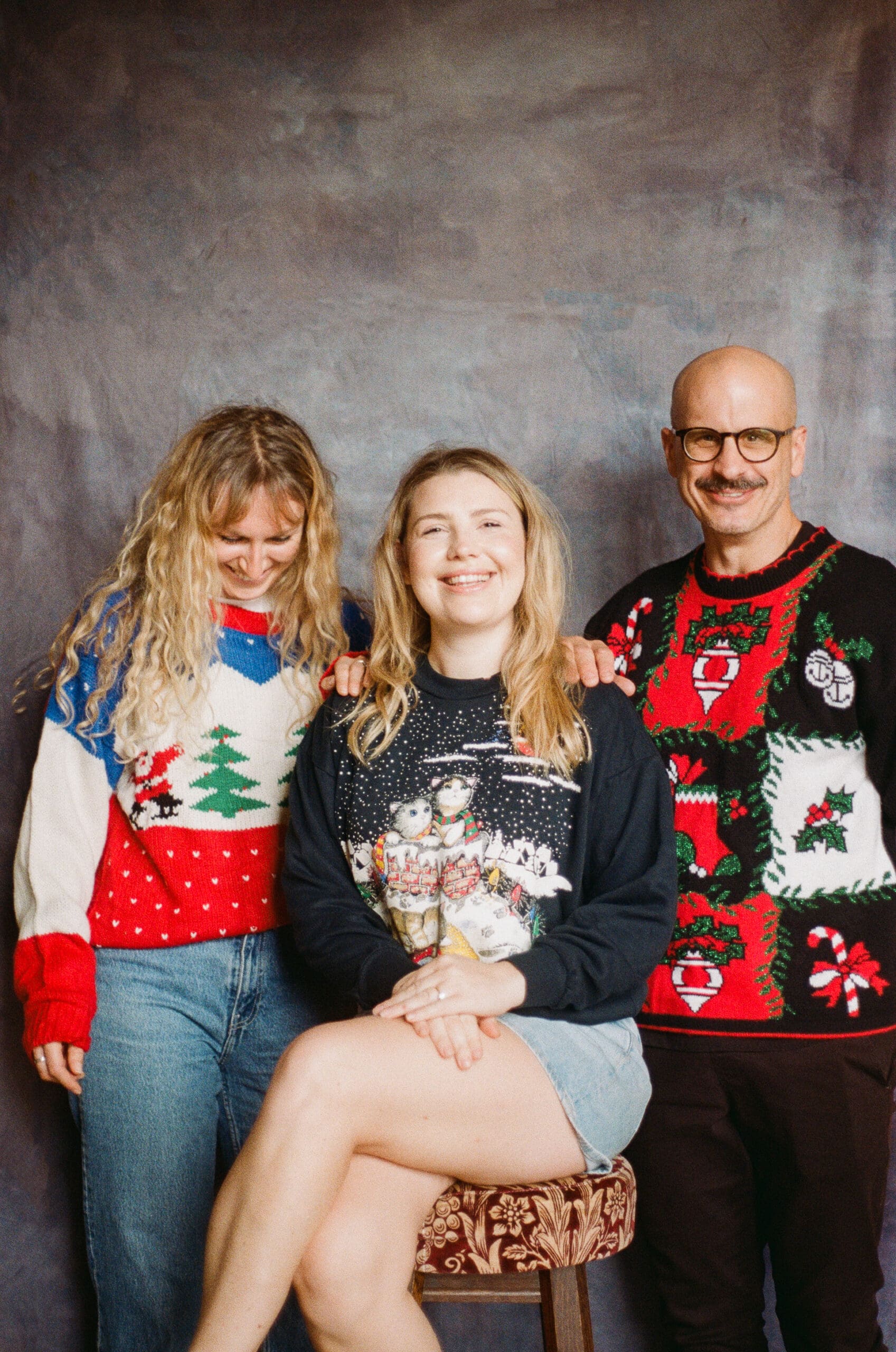 Three adults in festive Christmas sweaters pose for a photo; one woman sits on a stool smiling, while a man and another woman stand beside her, both smiling as well, against a plain studio backdrop.