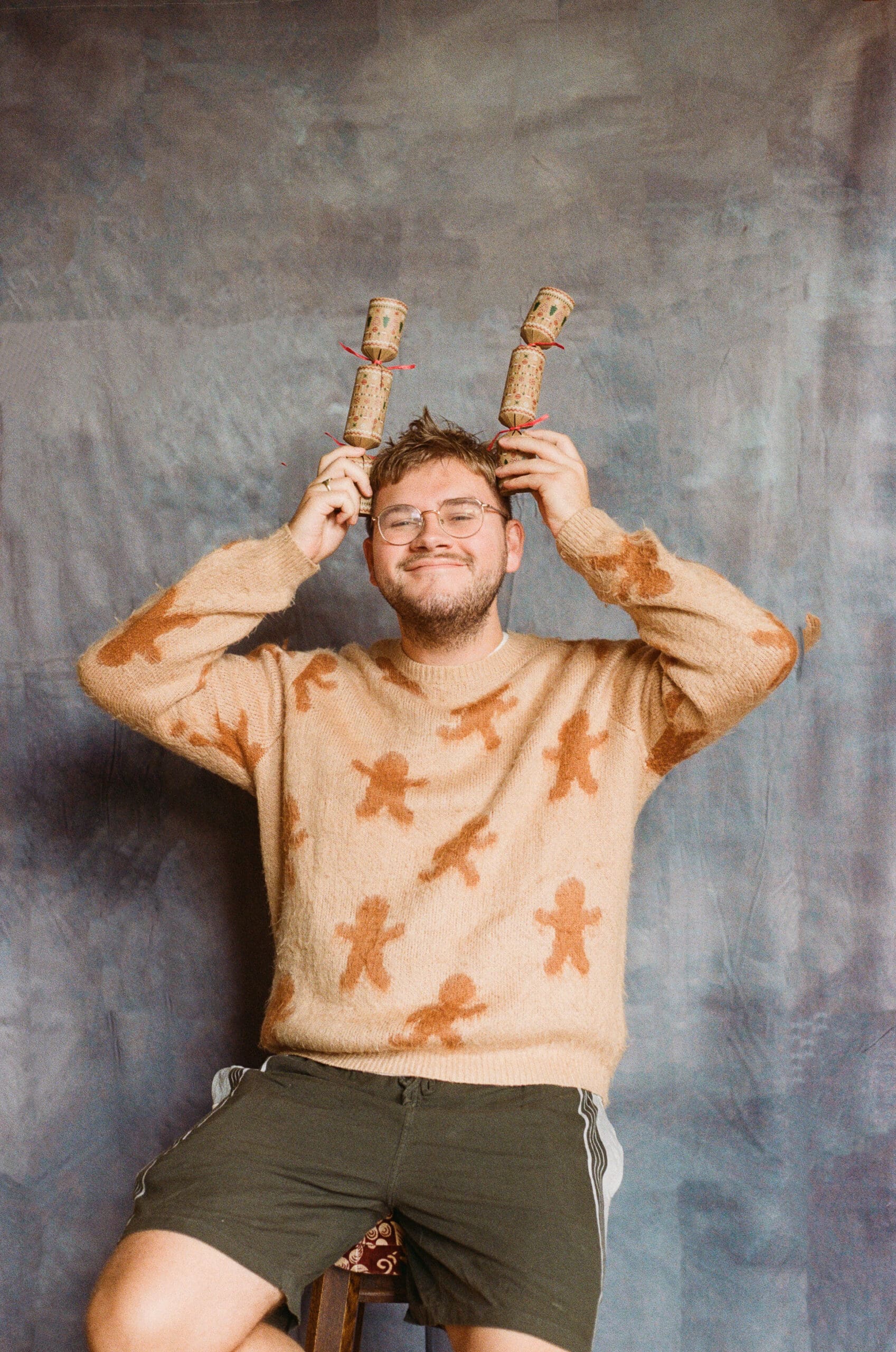 A man wearing glasses and a beige Christmas sweater with gingerbread figures sits on a stool, smiling with his eyes closed while playfully holding two festive crackers above his head like antlers.
