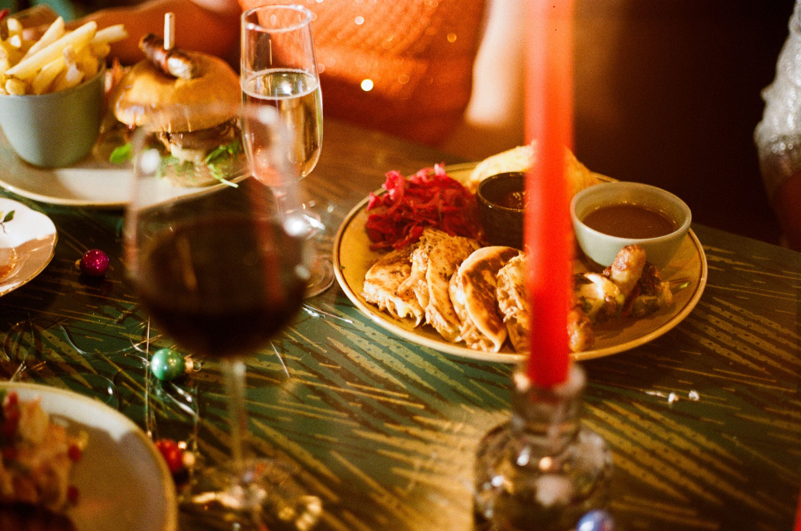 A festive Christmas dinner table with plates of food including a burger, fries, roast meat, and sides. Glasses of wine and champagne sparkle beside a lit red candle in the foreground, as people gather around to celebrate.