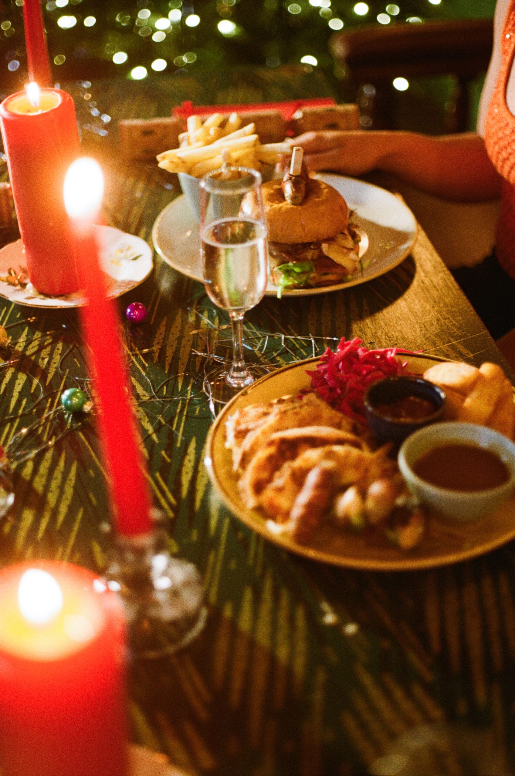 A festive Christmas table setting with lit red candles, a plate of holiday food including roasted vegetables and gravy, a burger with fries, a glass of sparkling wine, and holiday decorations in the background.