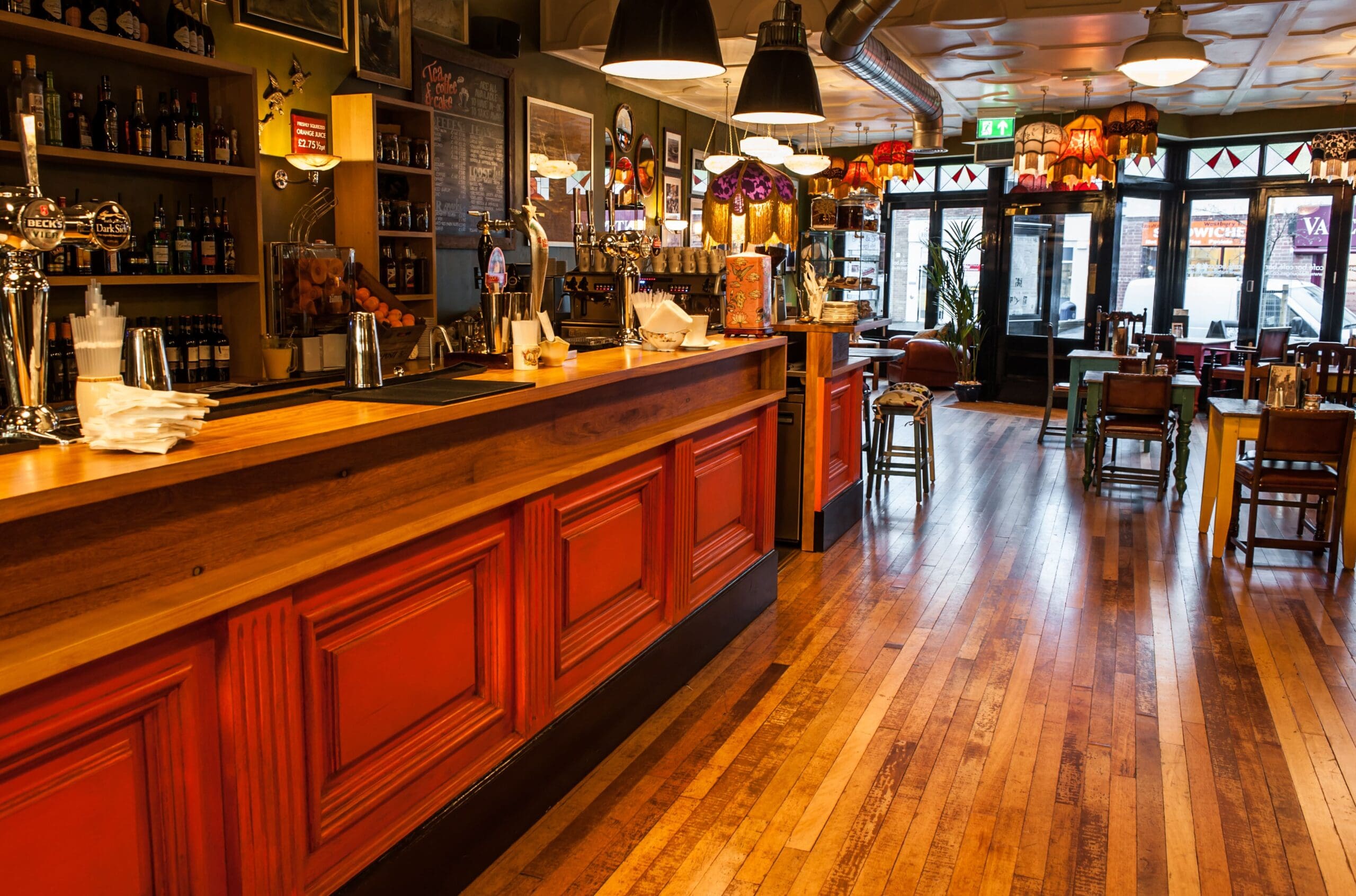 Cozy bar interior with a long wooden counter, red paneling, and wooden floors. Hanging lamps and tables with chairs create a warm Grupo atmosphere. Shelves behind the bar hold bottles and glasses, while large windows let in natural light.