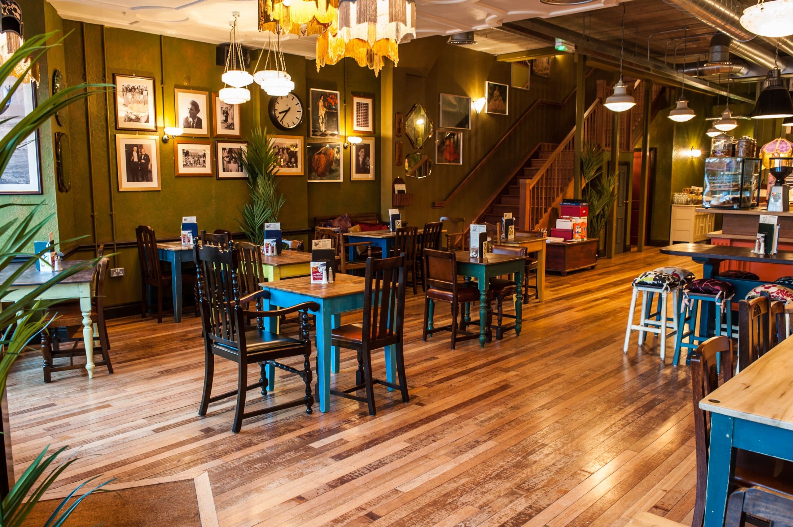 A cozy café interior with wooden floors, eclectic tables and chairs for any grupo, framed photos on green walls, a staircase, and warm lighting from hanging lamps create a welcoming atmosphere.