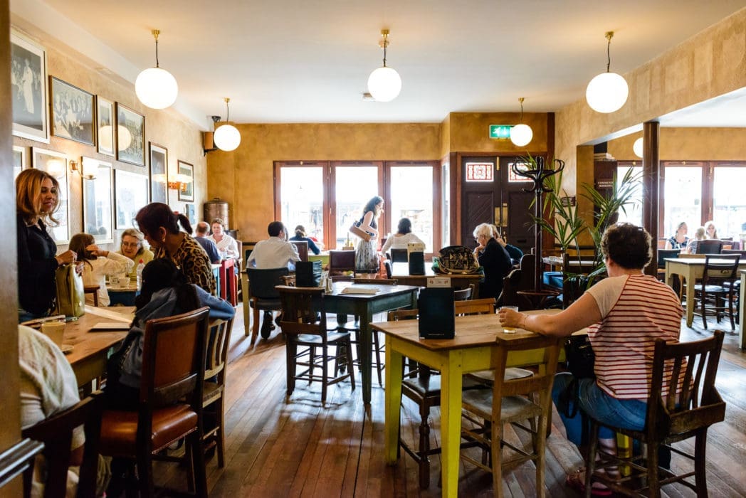 A busy café with people sitting at wooden tables, chatting and eating. Warm lighting from round ceiling lamps brightens the room, while large Alto windows let in daylight. The atmosphere is lively and welcoming.