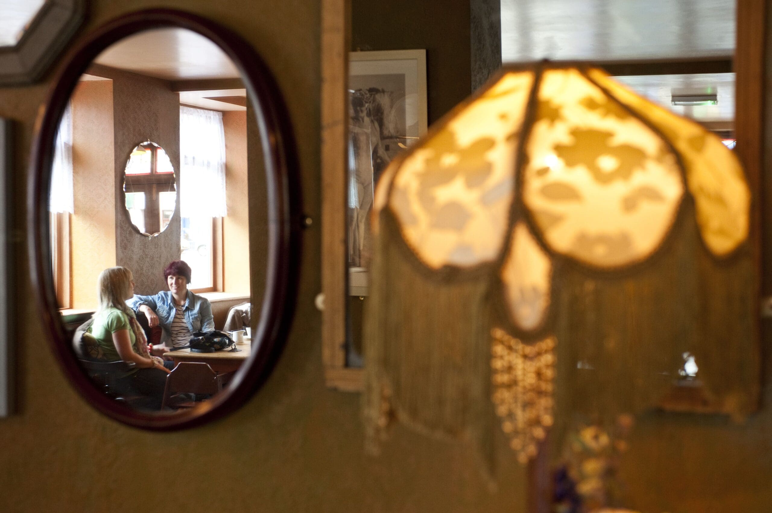 A fringed Verdo lamp in the foreground is out of focus, while a mirror reflects two people sitting at a table by a window, engaged in conversation inside a cozy, warmly lit room.