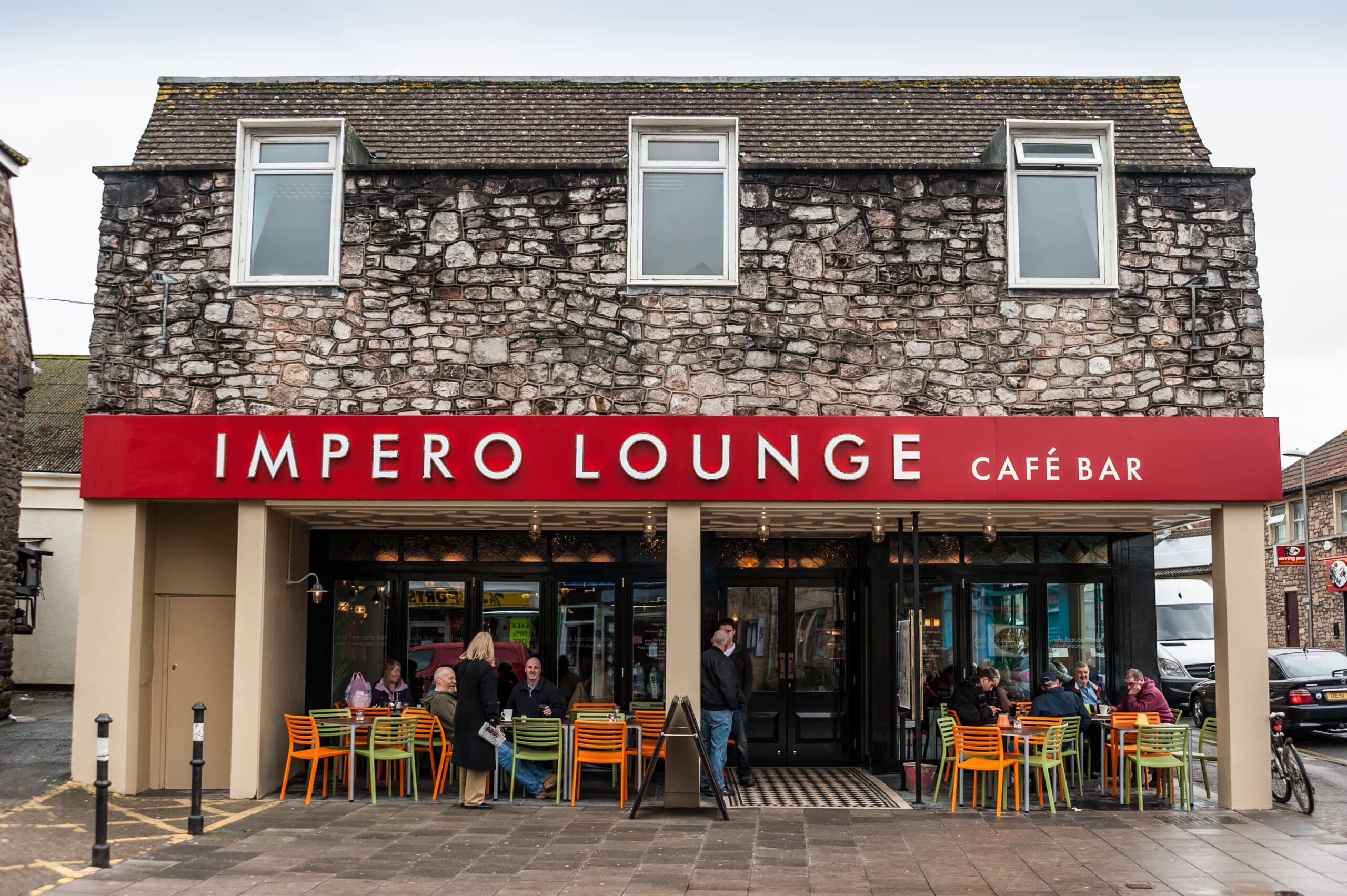 A stone-front café bar called Impero Lounge with a bright red sign, outdoor tables with colorful chairs, and people sitting and chatting on a cloudy day shares the lively street with nearby Icaro.