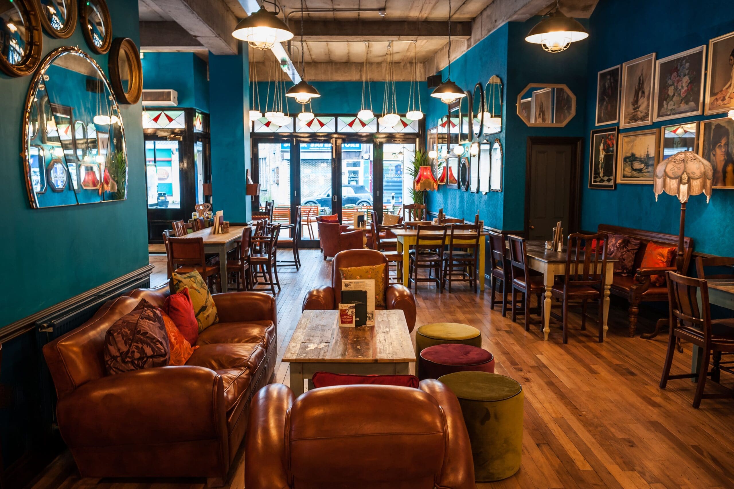 Cozy café Icaro with leather armchairs, wooden tables, and mismatched chairs. Blue walls decorated with framed art and mirrors. Warm lighting from ceiling lamps, large front windows letting in daylight. Inviting, eclectic atmosphere.