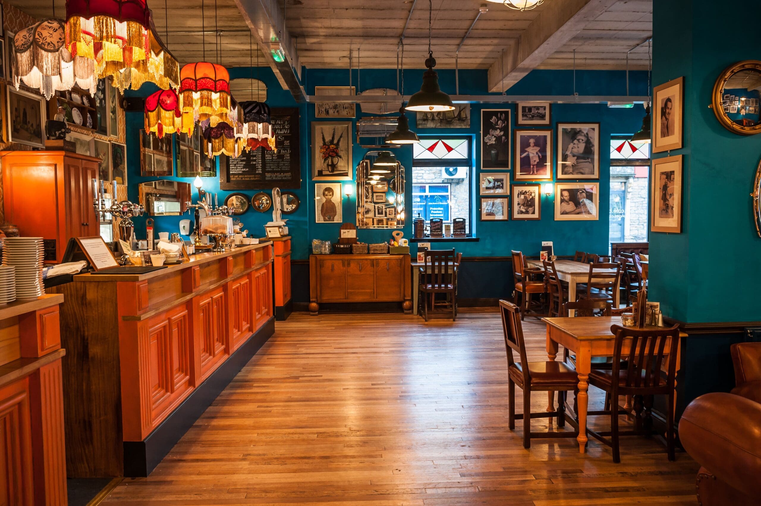 A cozy café with vintage decor, featuring wooden tables and chairs, red-shaded hanging lamps, blue walls adorned with framed photos, and a wooden counter along the left side—welcome to Icaro.