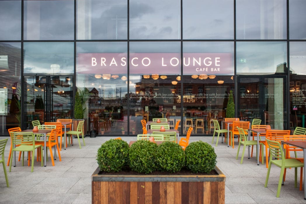 The Brasco Lounge café bar’s outdoor seating area features green and orange chairs, potted plants, and large glass windows offering a view into the cozy Brasco interior with warm lights and wooden furniture.