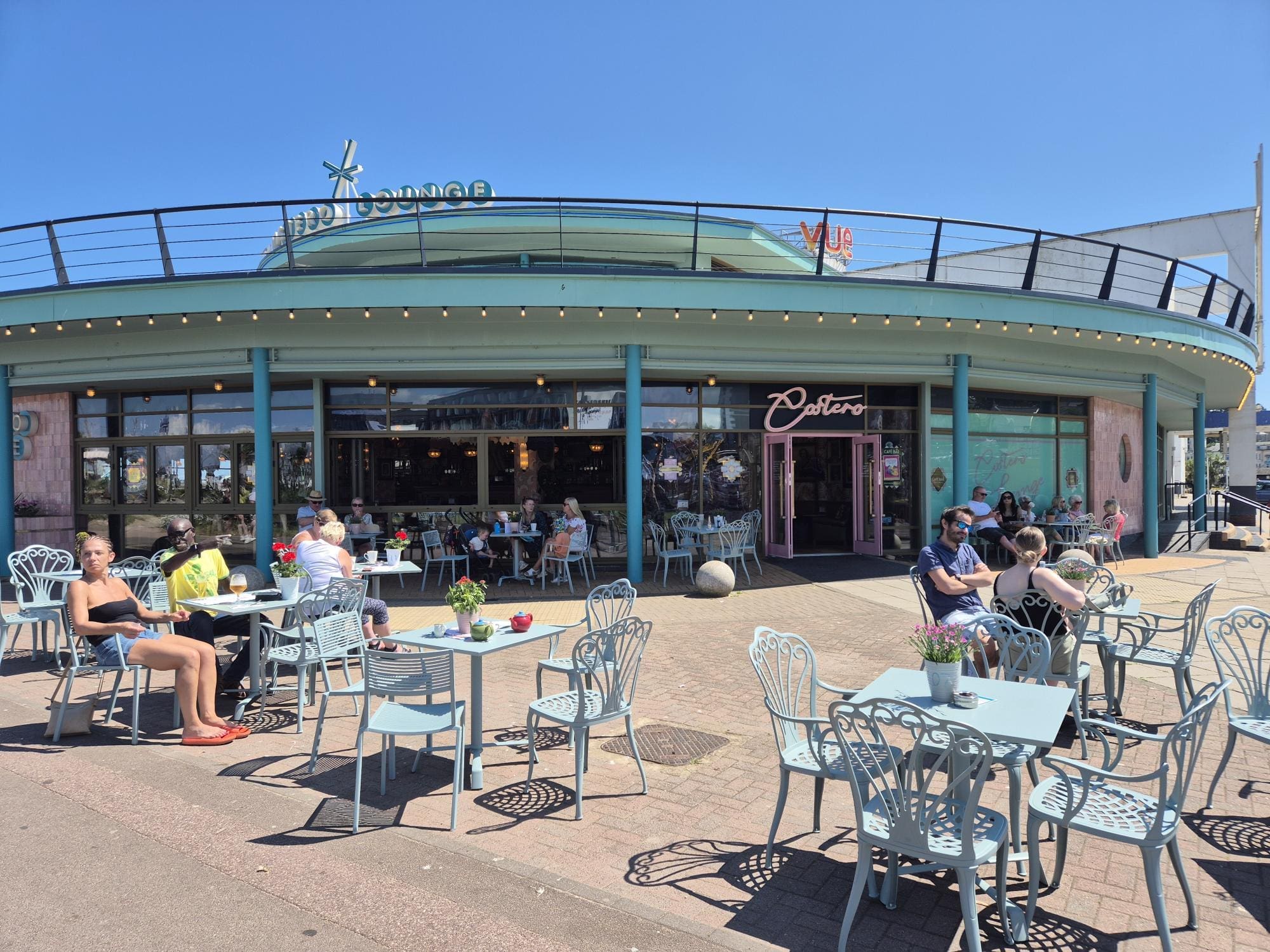 People sit at light blue tables and chairs outside Costero, a pastel-colored café with large windows on a sunny day. Some are eating or drinking, while others relax and chat under the clear blue sky.