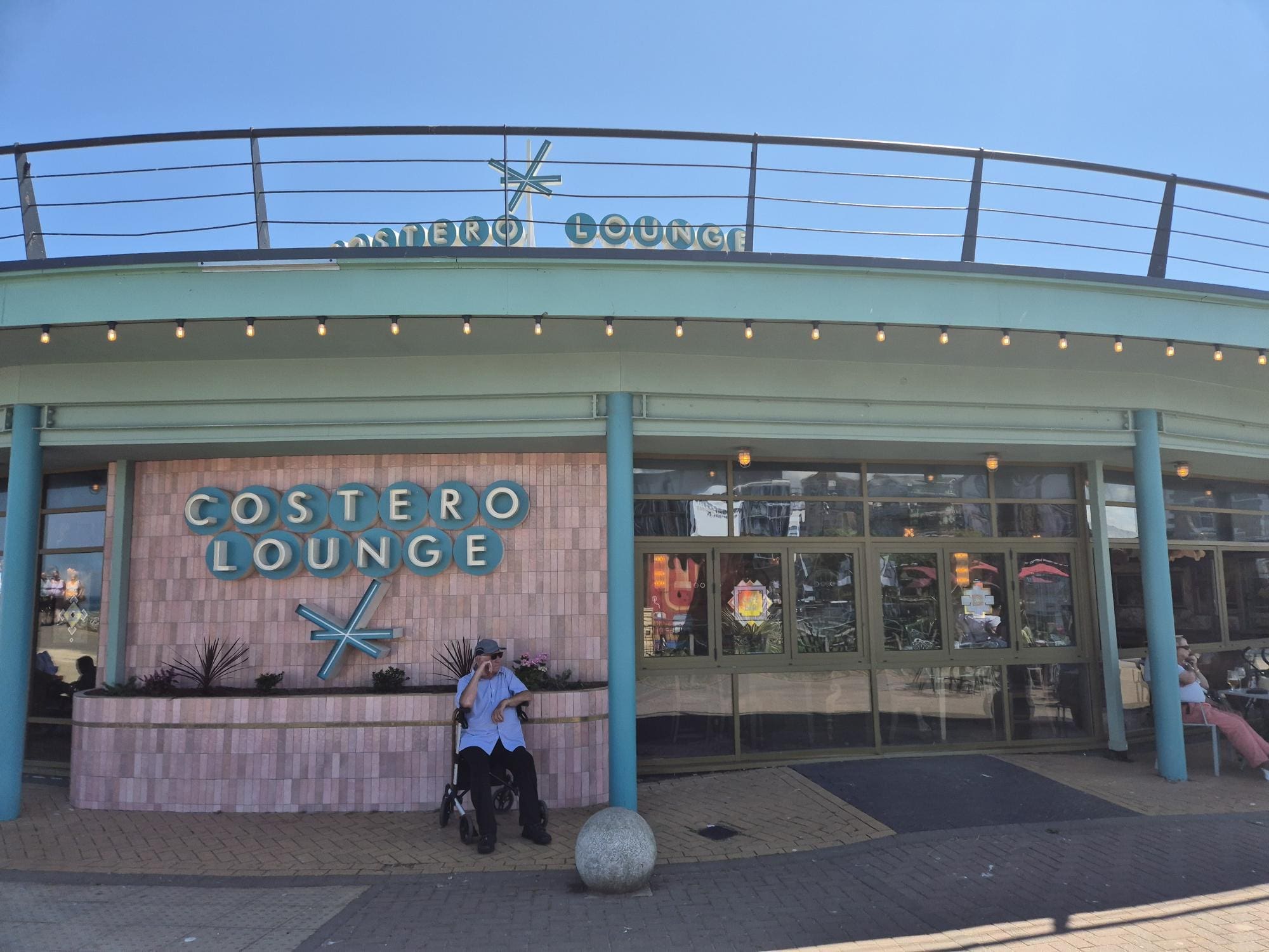 A person in a wheelchair sits outside Costero Lounge, a cafe with turquoise accents and large windows, basking in the sun. The "COSTERO LOUNGE" sign stands out above the entrance.