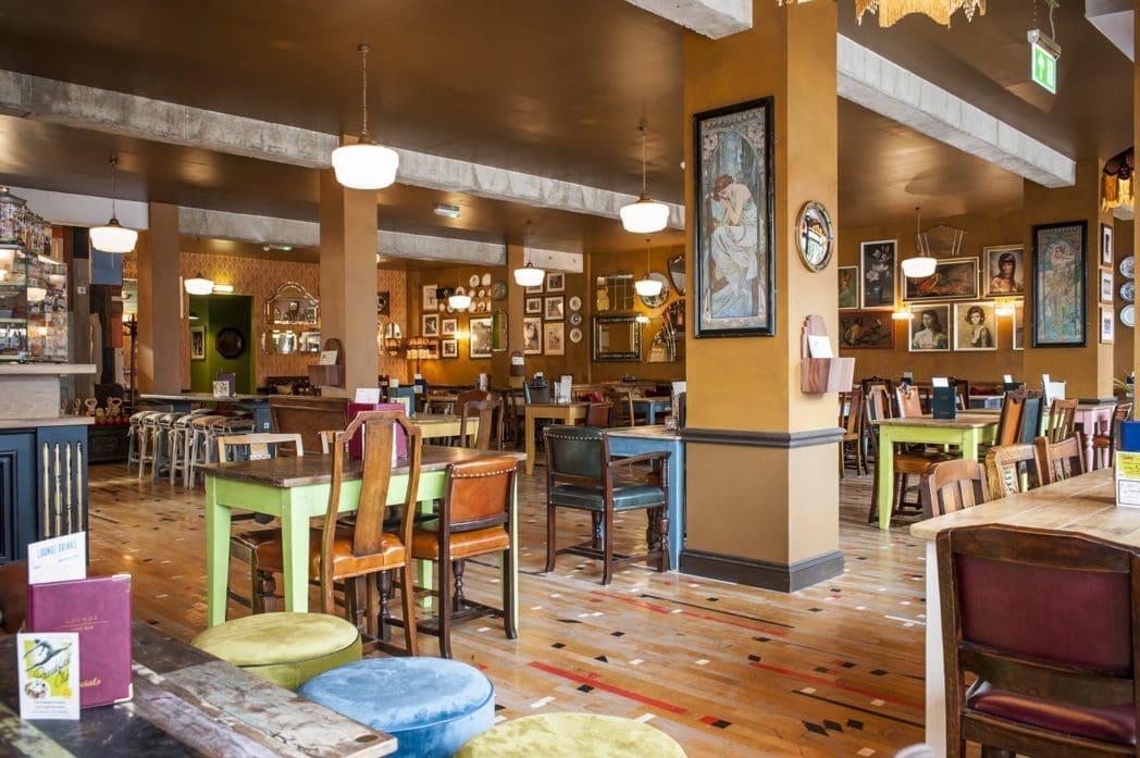 A cozy, eclectic café with colorful mismatched chairs, wooden tables, framed art on mustard walls, pendant lights, and a polished wooden floor—Bosco creates a warm and inviting atmosphere.