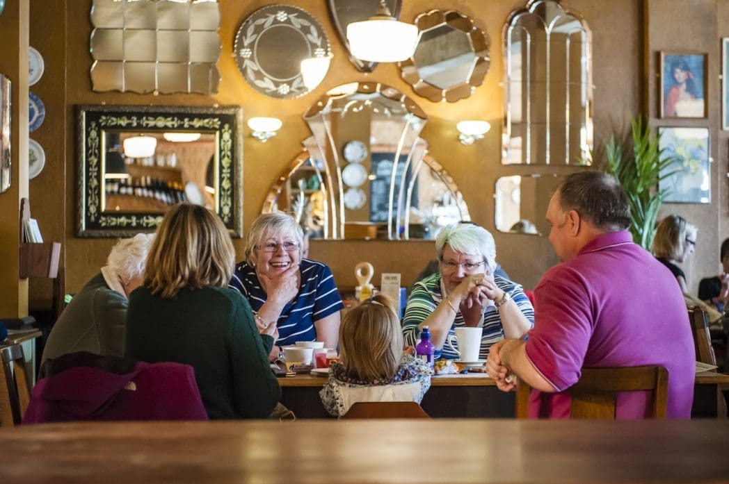 A group of older adults and a young child sit around a table in cozy Bosco café, chatting and smiling. Decorative mirrors and framed art hang on the wall behind them, reflecting the warm, inviting atmosphere.