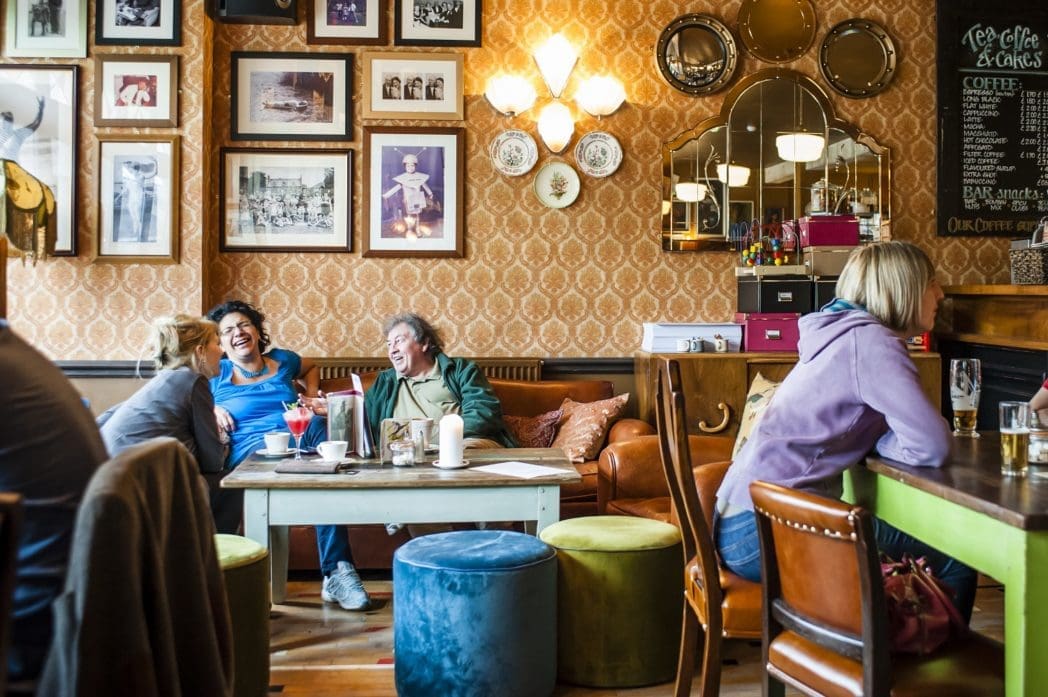 Three people sit and laugh on a couch in Bosco, a cozy café with eclectic décor, framed photos on the wall, colorful poufs, and a chalkboard menu. Another person sits alone at a nearby table facing away.