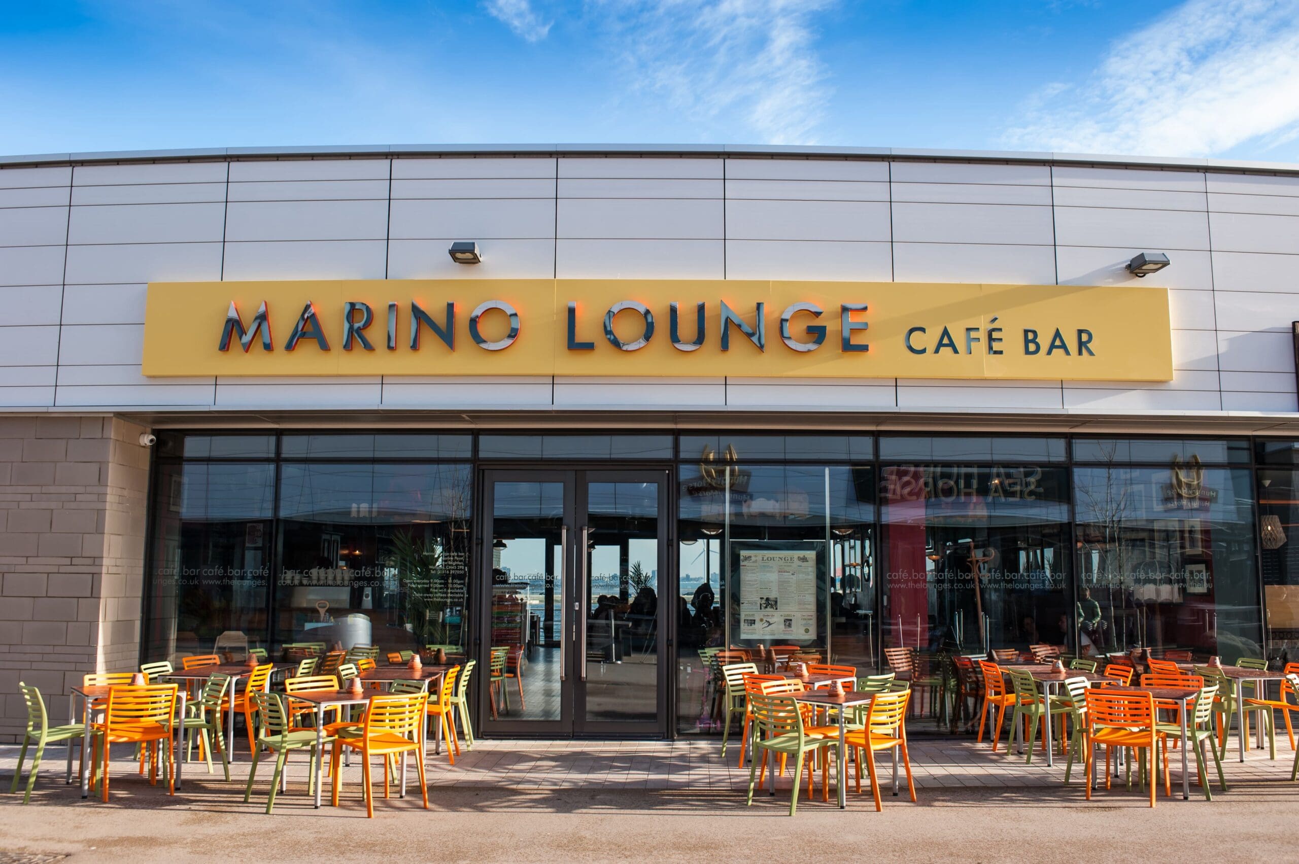 The exterior of Marino Lounge Café Bar, with its yellow Marino sign above glass doors and windows, features colorful orange, yellow, and green chairs on the patio under a blue sky.