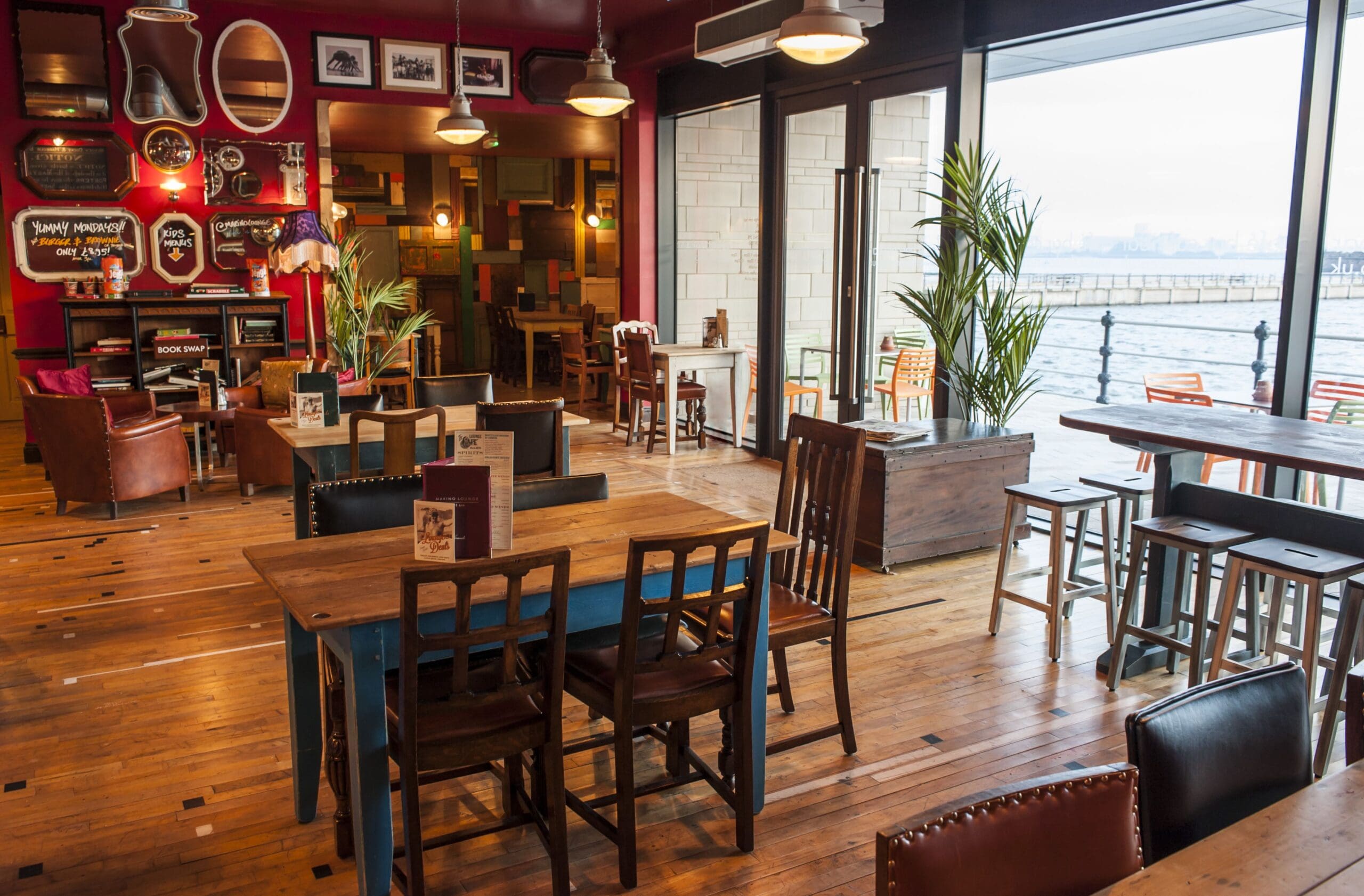 A cozy café with wooden floors, mismatched tables and chairs, warm lighting, quirky wall decor, and large windows offering a stunning Marino waterfront view dotted with boats and a distant city skyline.