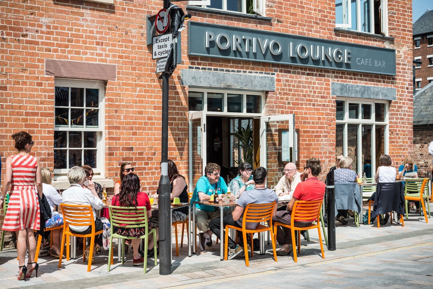 People relax at colorful chairs and tables outside Portivo Lounge on a sunny day, enjoying food and drinks beside the brick building. The café’s name, Portivo, is visible near large windows and a signpost in the background.