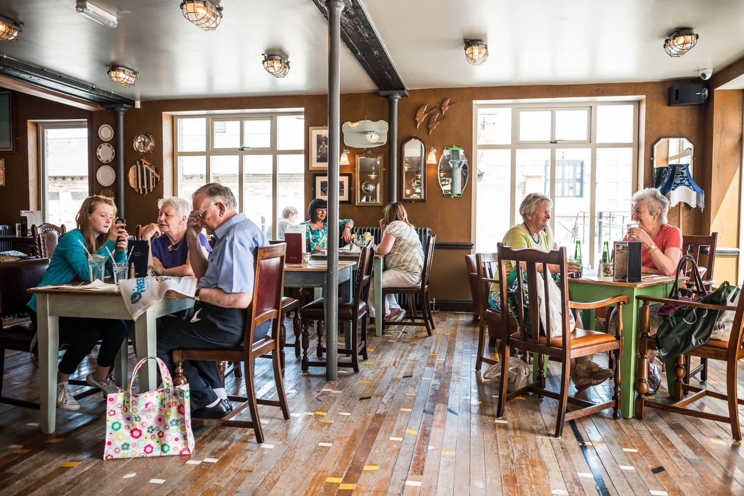 People sit at tables in cozy Portivo café, talking and eating. Sunlight streams through large windows, while pictures, mirrors, and wall art decorate the space. The wooden floor adds to the café’s warm, inviting atmosphere.