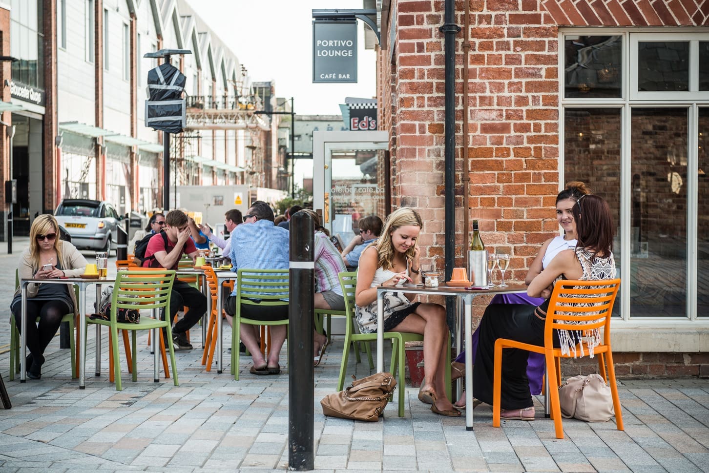 People are sitting at colorful tables and chairs outside Portivo café, chatting, eating, and using their phones. The lively scene features a brick building and café signs in the background.
