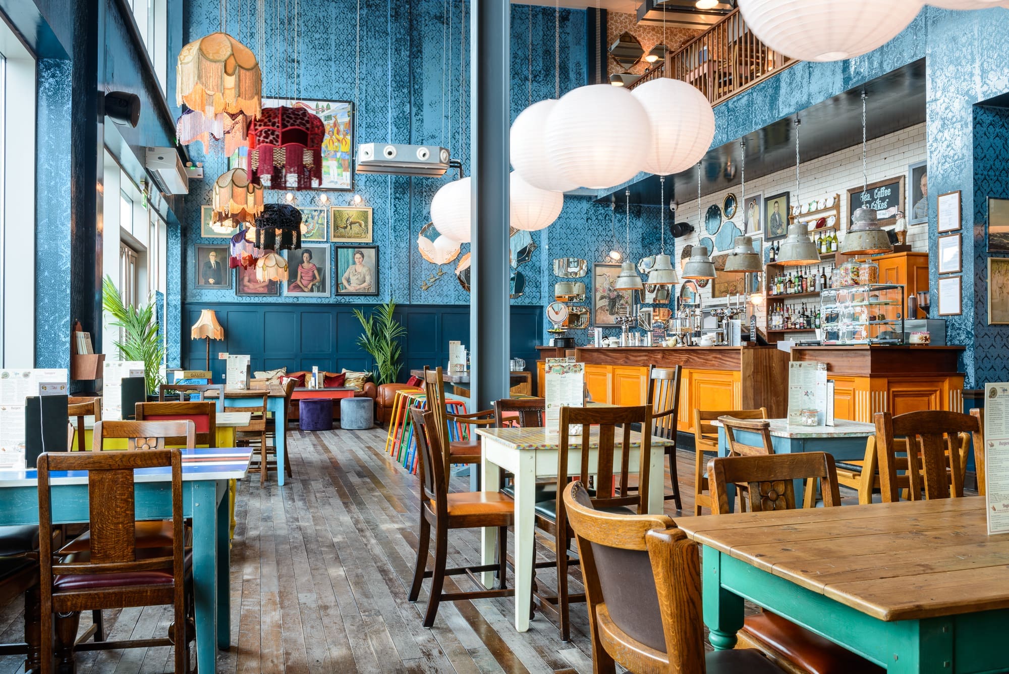 A colorful, eclectic café called Desco with mismatched wooden chairs, vintage decor, hanging lanterns, framed art on blue-patterned walls, and a long wooden bar counter. Large windows flood the space with natural light.