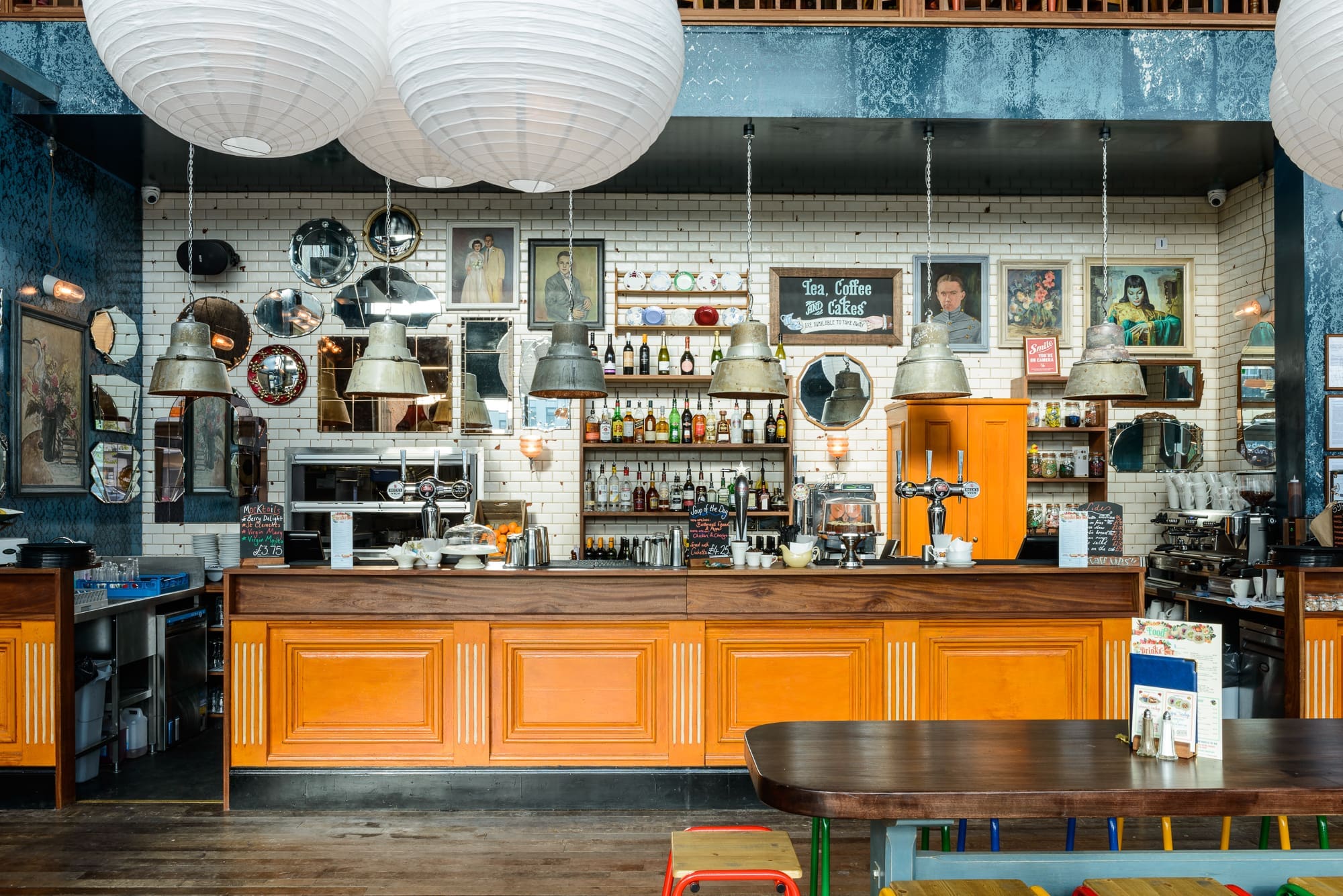 A stylish café interior by Desco with an orange wooden counter, hanging industrial lights, round paper lanterns, vintage mirrors, framed art, bottles on shelves, and wooden tables with colorful stools.