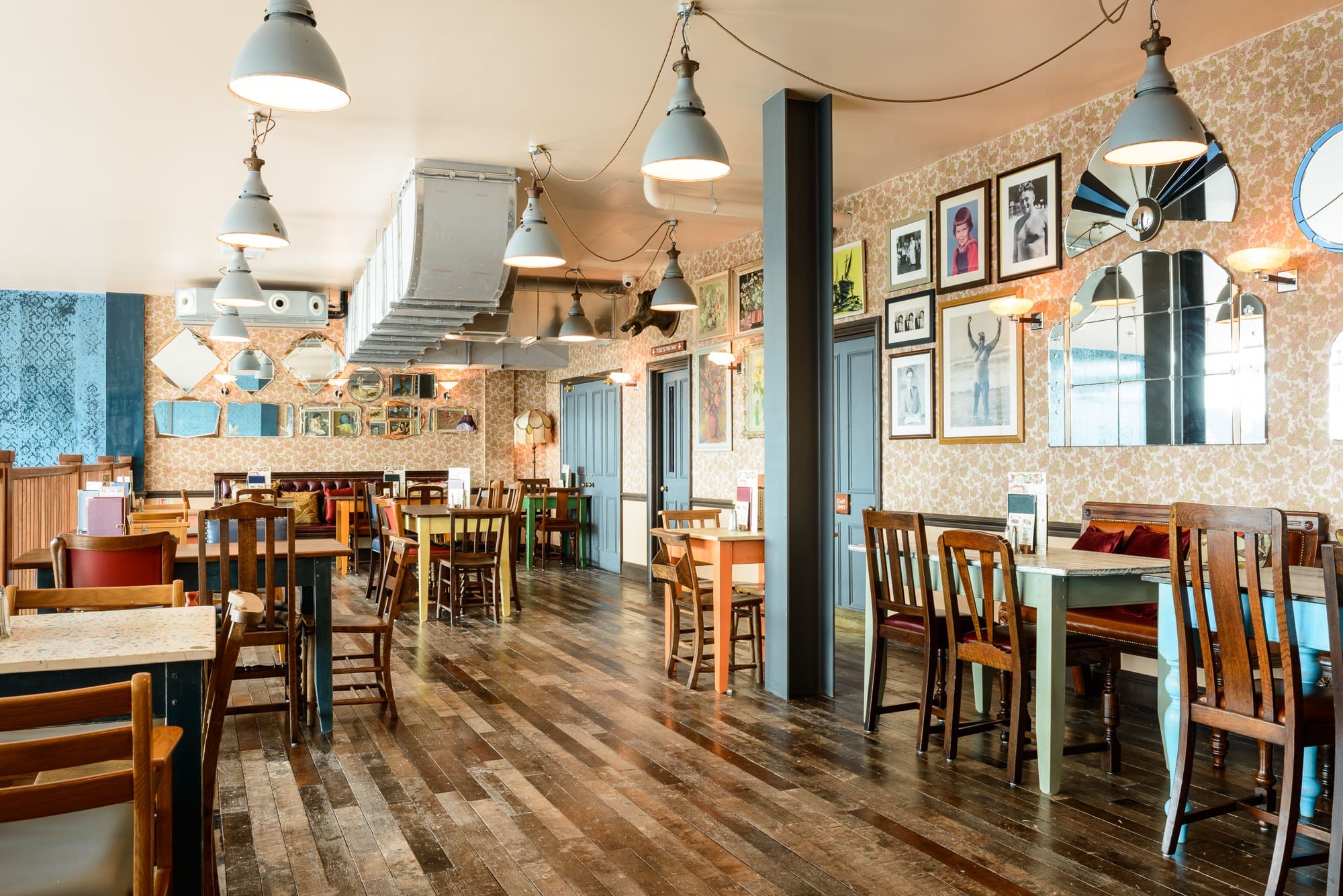 A cozy, vintage-style Desco restaurant interior with wooden floors, mismatched chairs and tables, patterned wallpaper, hanging lights, and framed pictures on the walls. The space is brightly lit and empty of people.