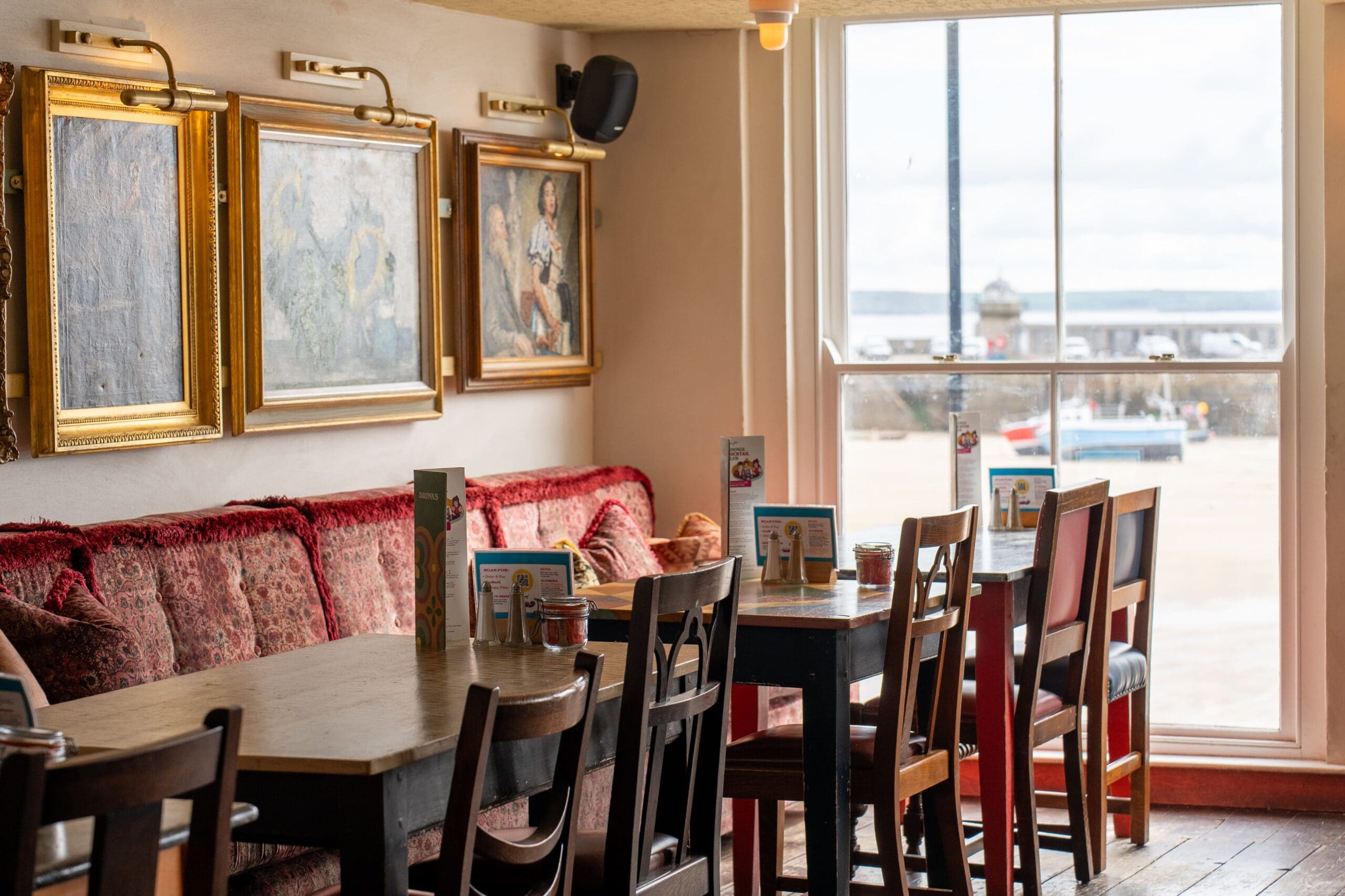 A cozy Tretho cafe interior with wooden tables and mismatched chairs beside a window overlooking a harbor. Framed paintings hang on the wall above a red cushioned bench, and menus are neatly arranged on the tables.