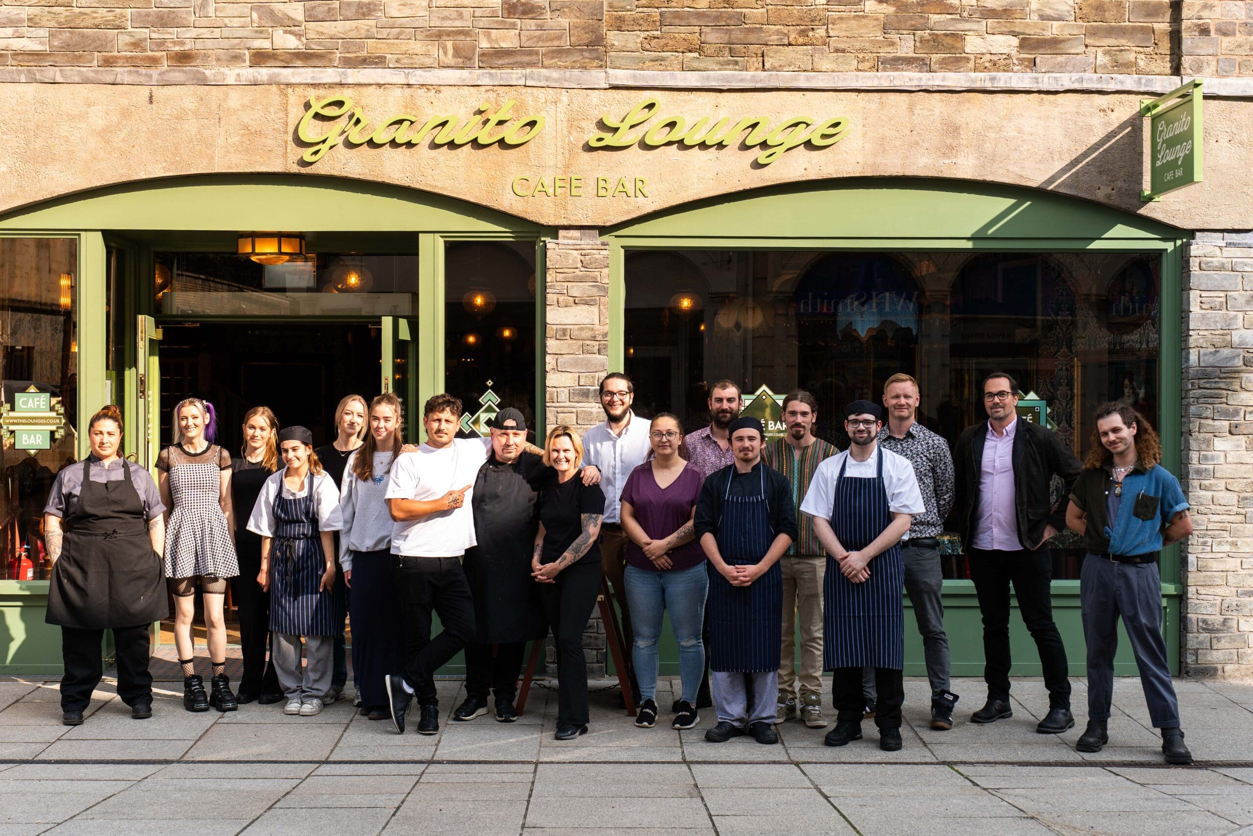 A group of sixteen people, including chefs and staff, stand smiling in front of Granito Lounge Café Bar on a sunny day. The café features distinctive granito green-framed windows and a sign above the entrance.