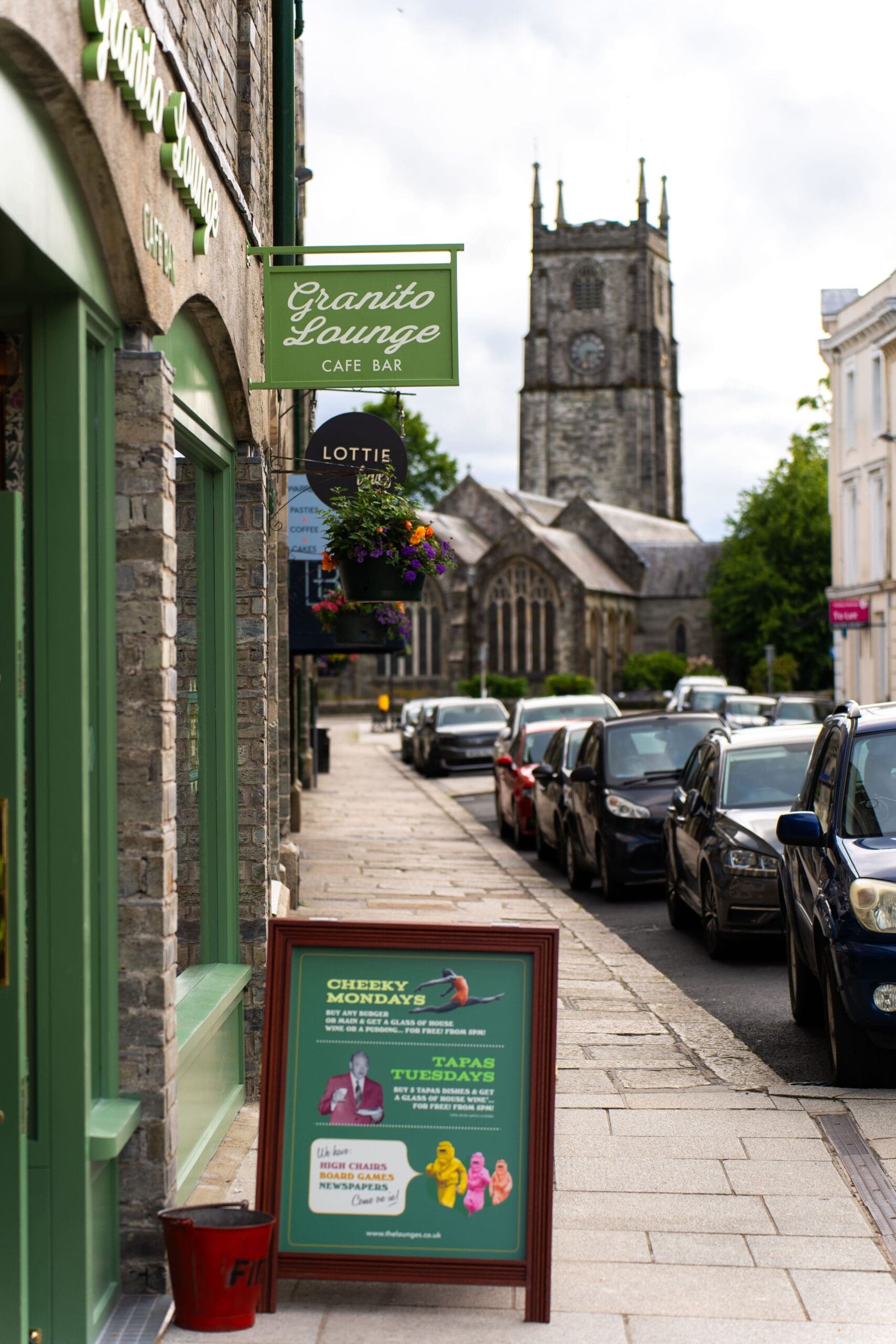 A street scene with parked cars, signs for Granito Lounge Café Bar and Lottie, hanging flower baskets, and a historic granito stone church with a clock and tall tower in the background.