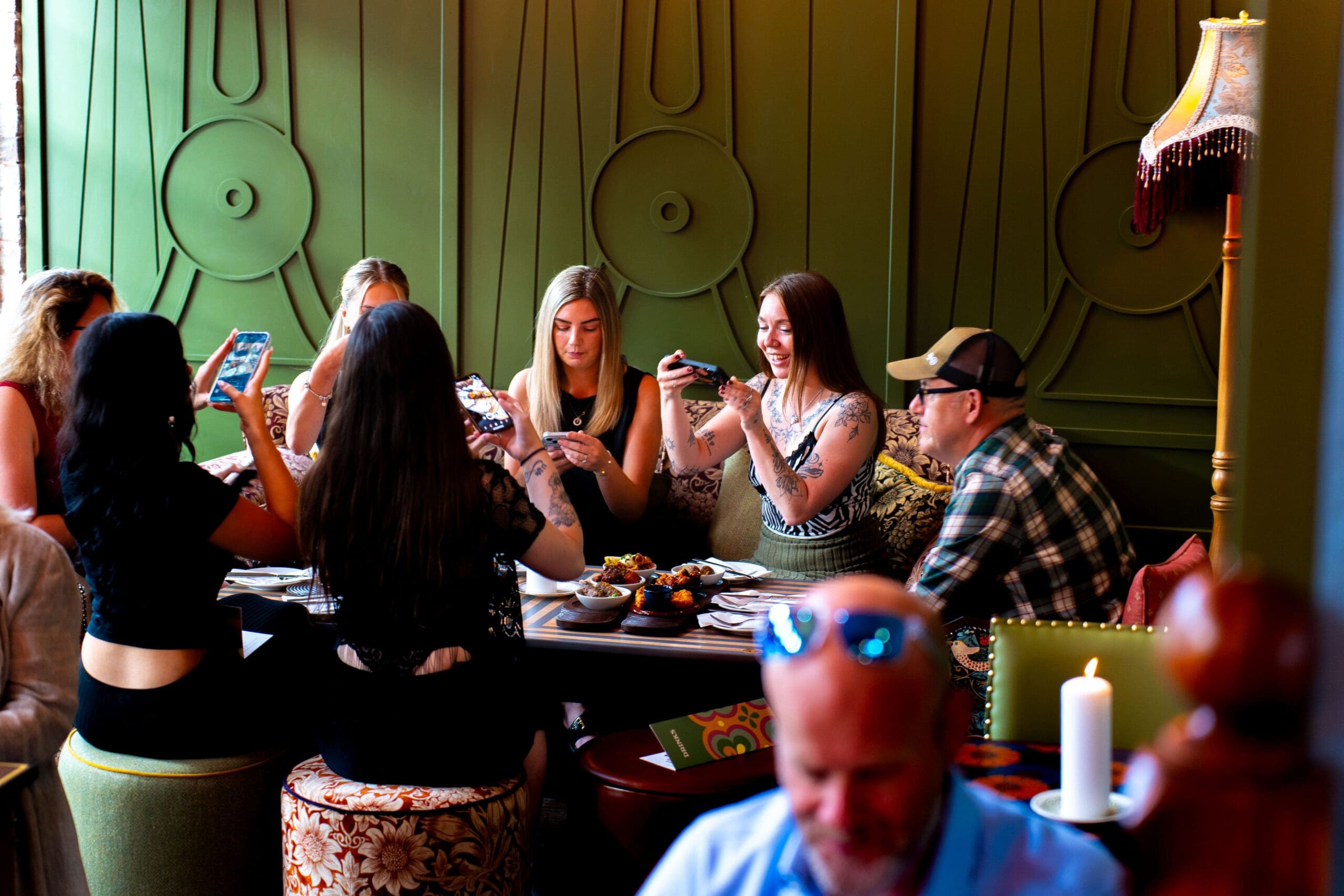 A group of women and one man sit around a restaurant table with a smooth granito surface, smiling and taking photos of their food. The background features a green wall with decorative designs and a vintage lamp.