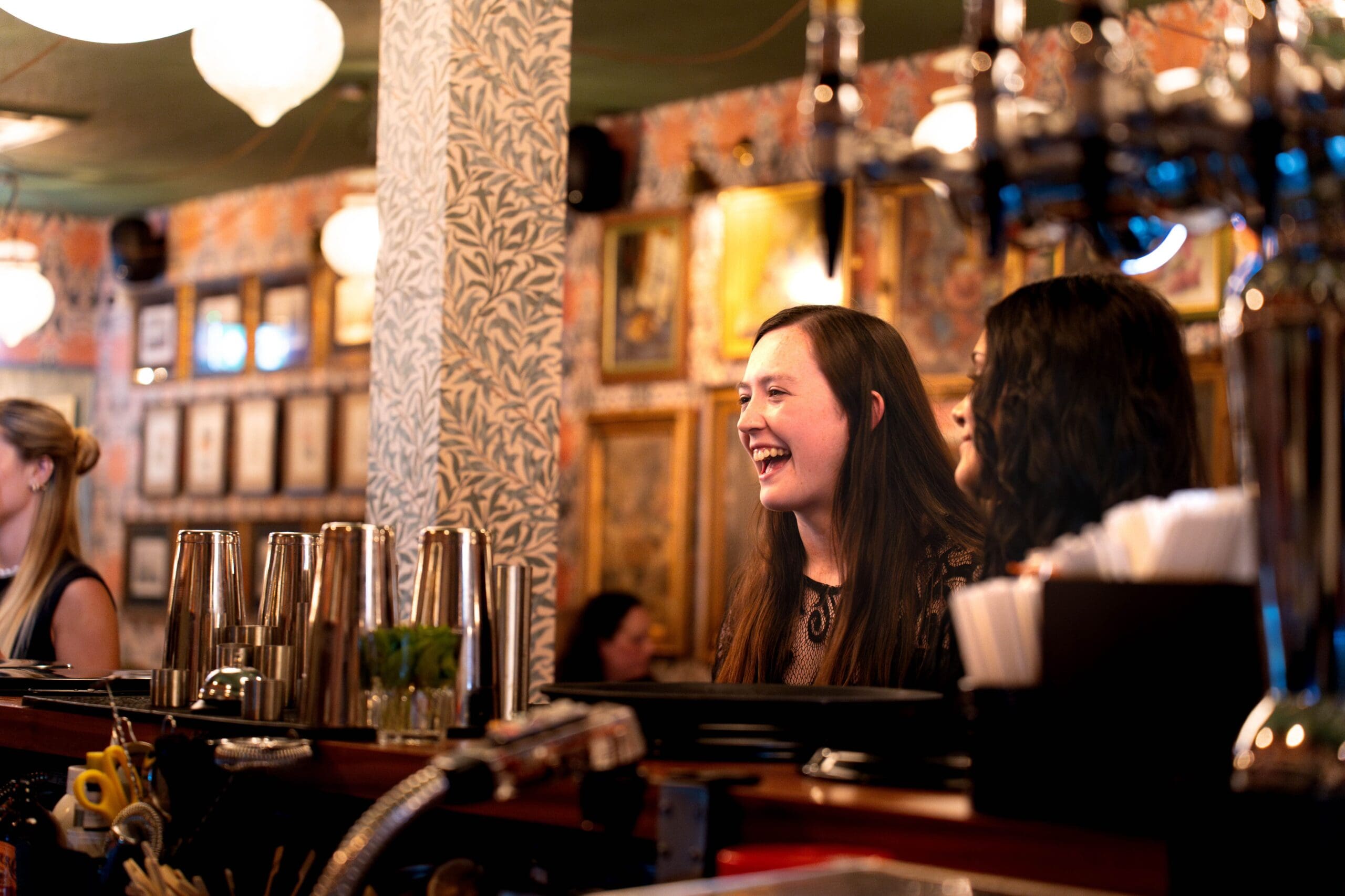 Two women stand behind a granito bar, smiling and talking, with cocktail shakers and bar tools in the foreground and framed pictures on the warmly lit wall behind them.