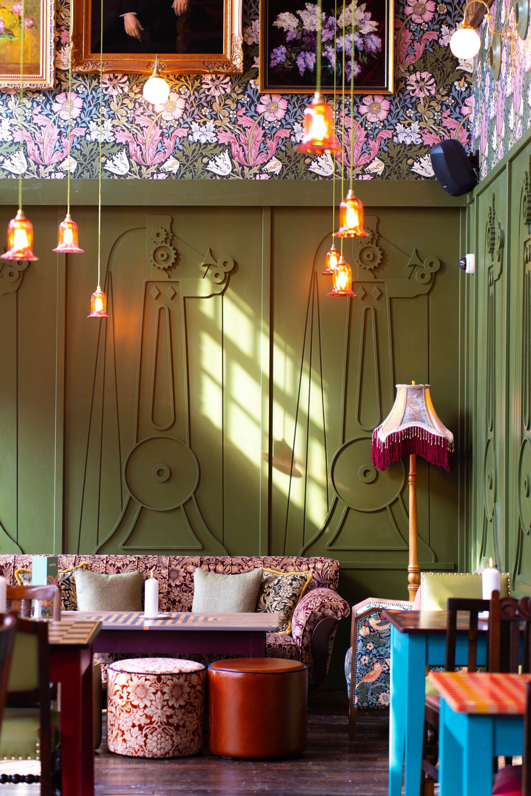 A cozy, vintage-style cafe with green paneled walls, patterned wallpaper, framed art, and hanging pendant lights. A floral sofa and granito-topped low tables sit near cushioned stools as sunlight casts shadows on the wall.