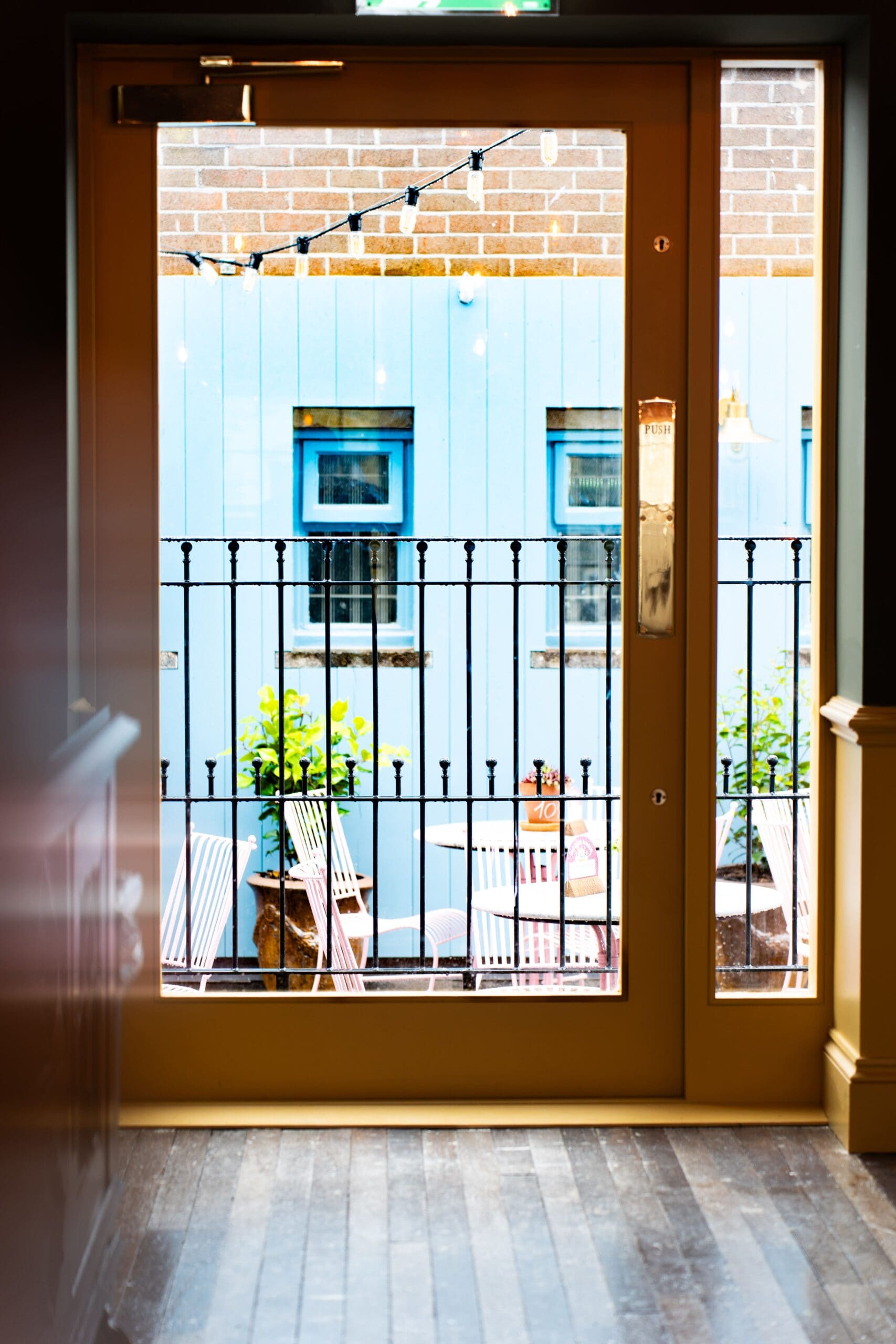 View through a glass door onto a small patio with striped deck chairs on a smooth granito floor, potted plants, string lights, and a light blue wall with windows in the background. The scene is bright and inviting.