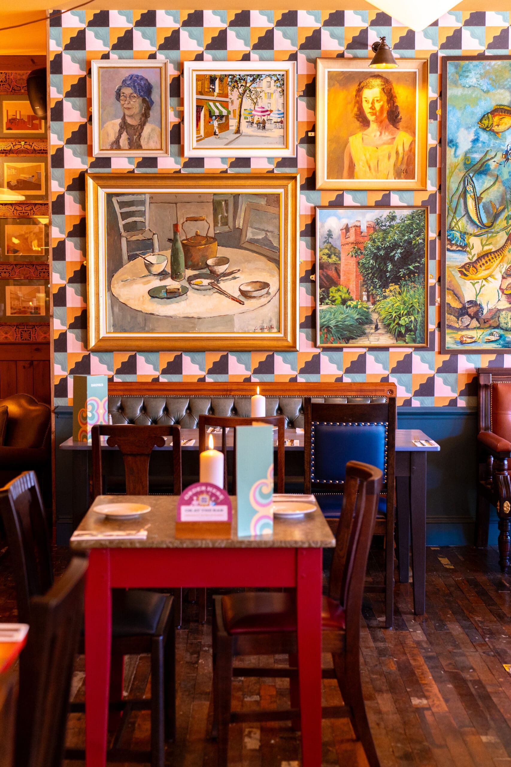 A cozy restaurant interior with wooden tables and chairs, candles on the tables, and a wall covered in divino colorful paintings and patterned wallpaper.