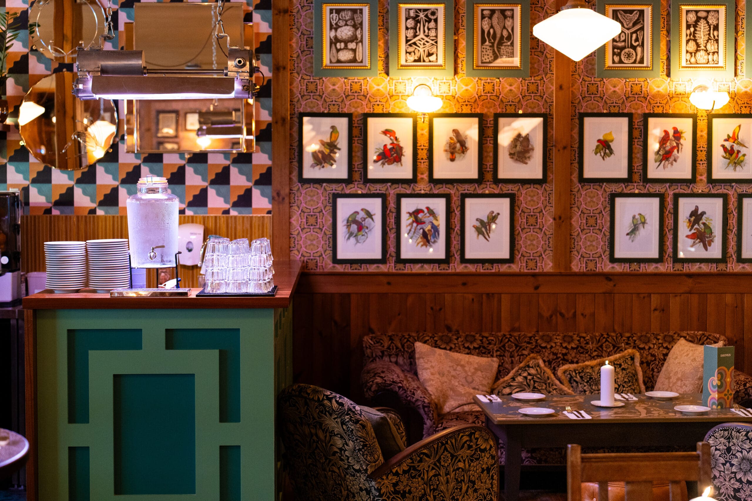 Cozy restaurant interior with patterned walls, framed bird artwork, cushioned seating, and a small dining table set with napkins. A green counter holds glasses, plates, and a large water dispenser. Divino warm ambient lighting sets the mood.