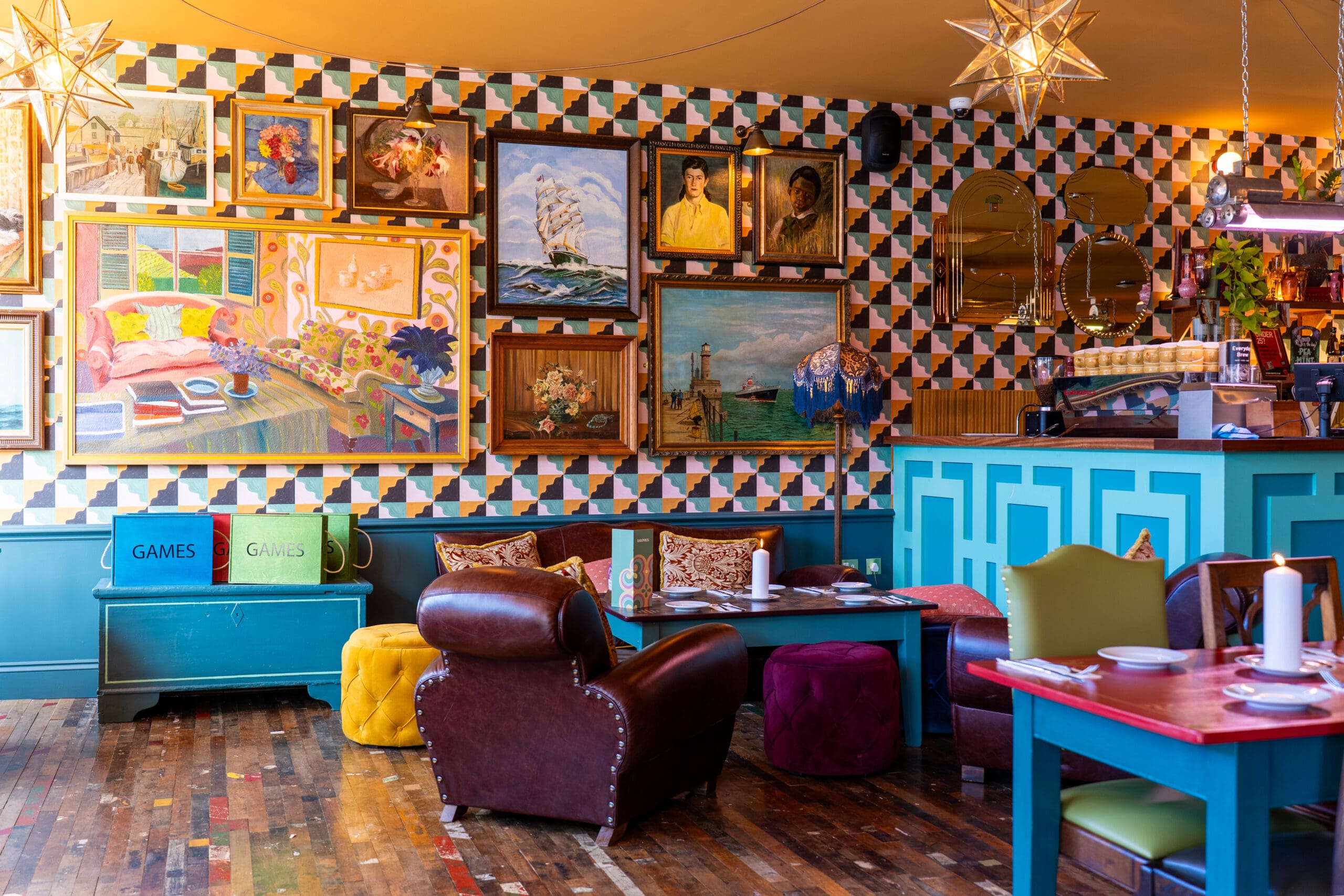Colorful cafe interior with eclectic furniture, star-shaped hanging lights, patterned wallpaper, and a divino gallery wall of assorted paintings. The space features tables, armchairs, and a blue counter with coffee equipment.