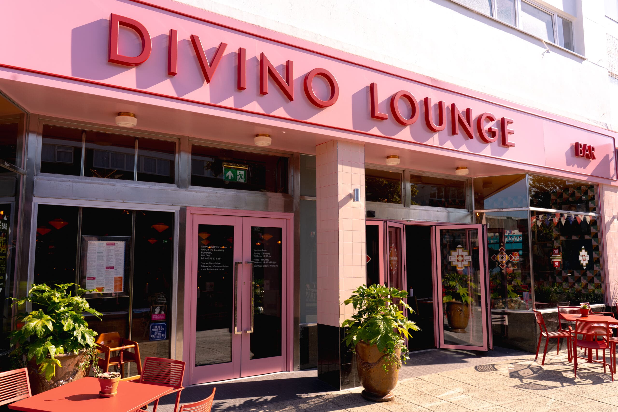 Exterior of Divino Lounge bar with vibrant pink signage, large glass doors, potted plants, and red outdoor tables and chairs on a sunny day. The welcoming divino entrance offers a glimpse of the stylish interior decor.