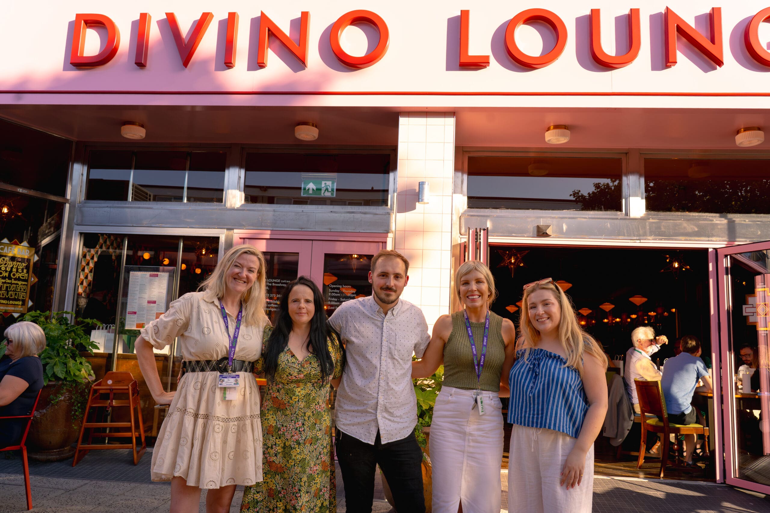 Five people stand smiling in front of the brightly lit entrance to Divino Lounge on a sunny day. The group appears happy and relaxed, with several guests enjoying their meals inside Divino’s inviting restaurant behind them.