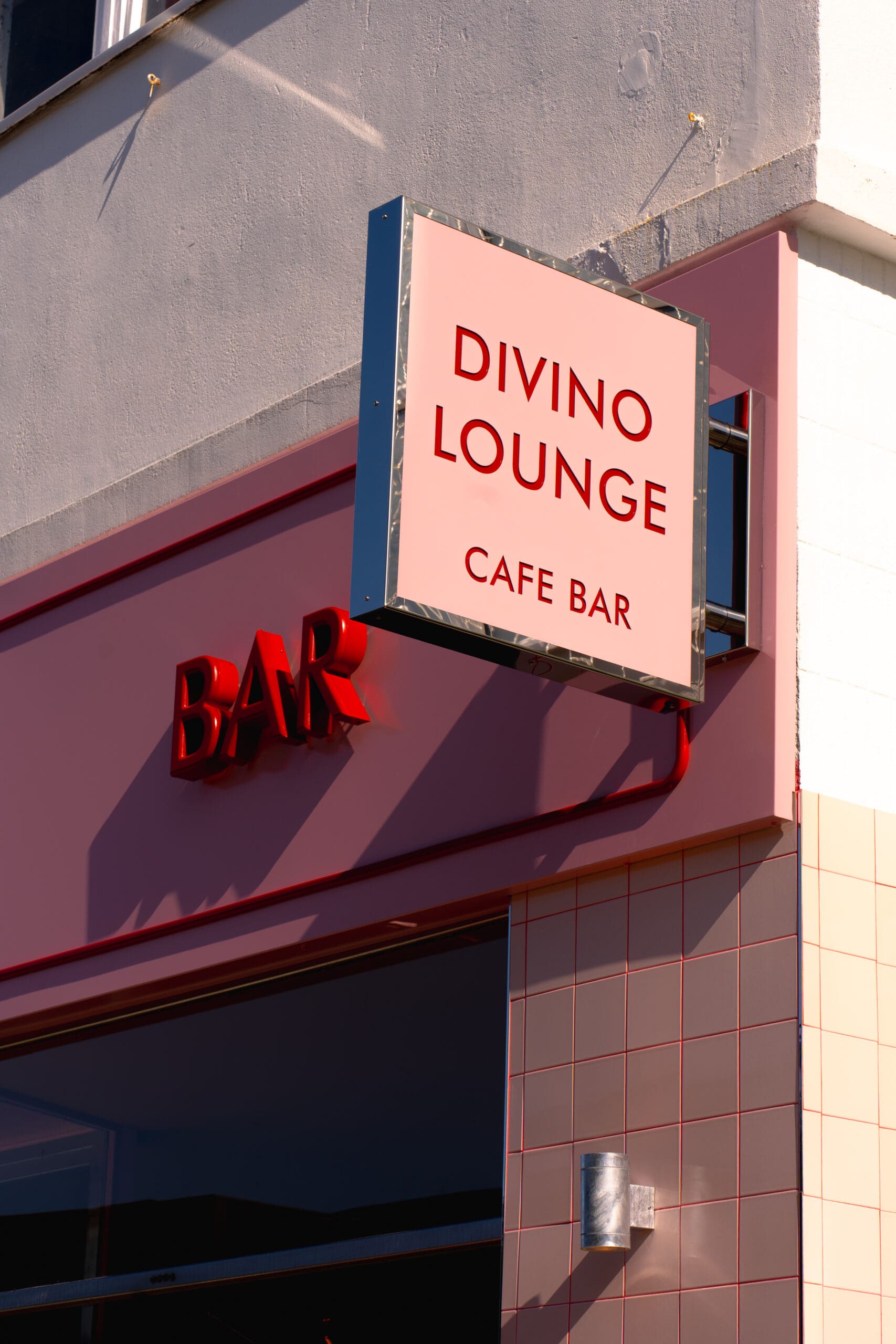 A modern café bar exterior with a pink and white "DIVINO LOUNGE CAFE BAR" sign and a red illuminated "BAR" sign glowing on the pink-tiled wall below, creating a welcoming Divino vibe.