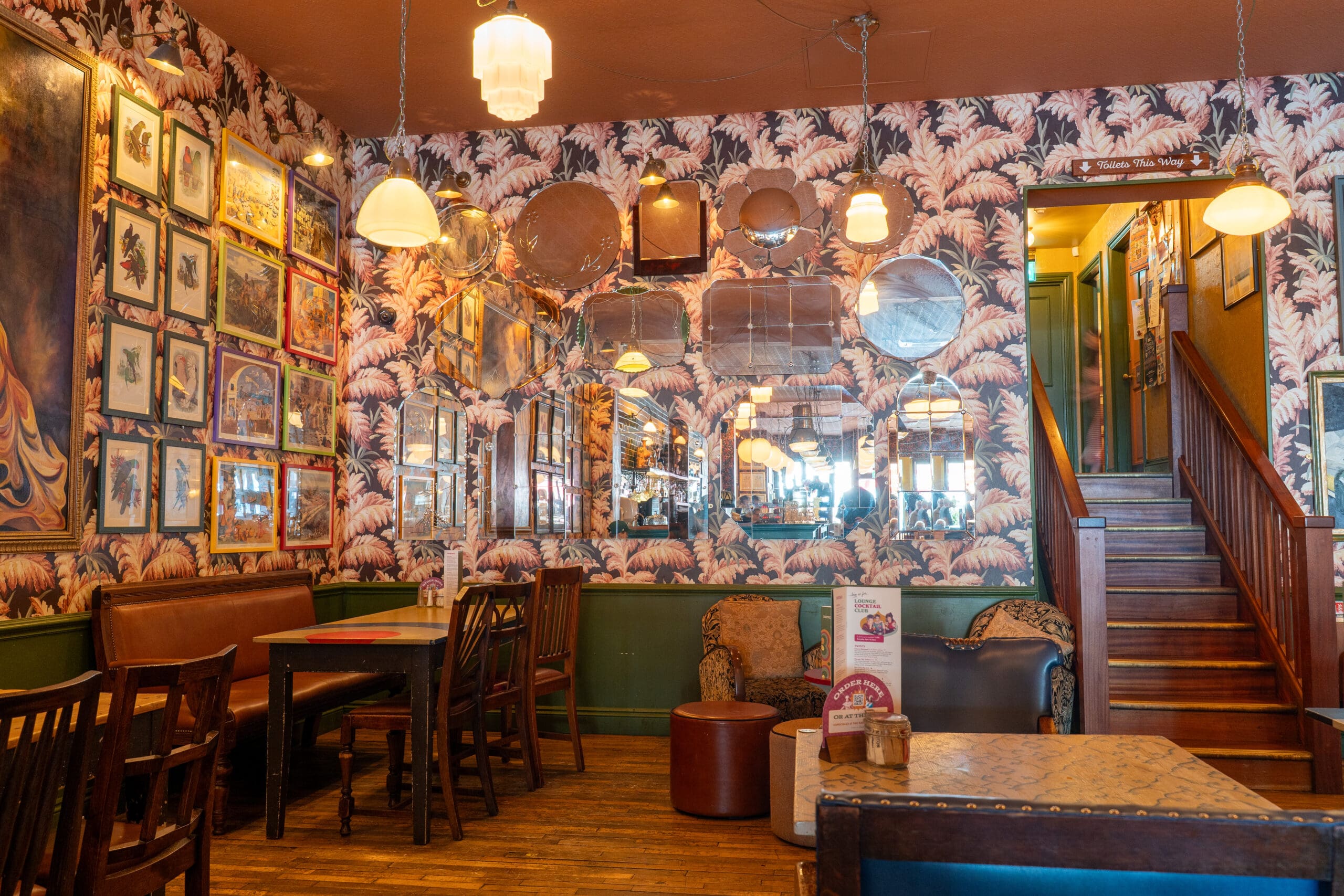 Cozy café interior in Porto with vintage-style wallpaper, assorted mirrors, framed art on the walls, wooden tables and chairs, upholstered seating, and a staircase leading to an upper floor. Warm lighting creates a welcoming atmosphere.