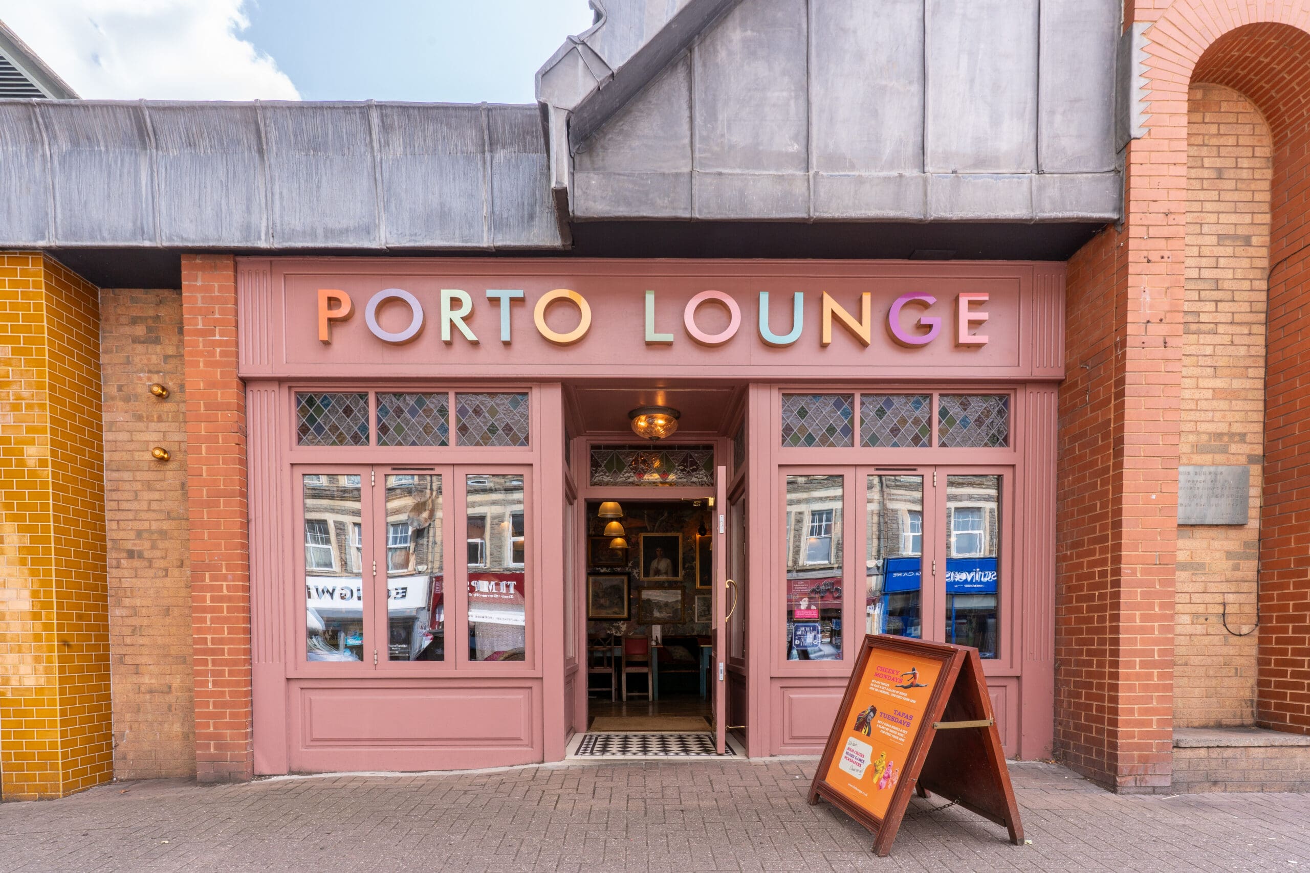 A pink Porto storefront with “PORTO LOUNGE” in colorful letters above the entrance. Large windows flank the door, and an A-frame sign stands on the sidewalk. The building features brick and metal detailing.