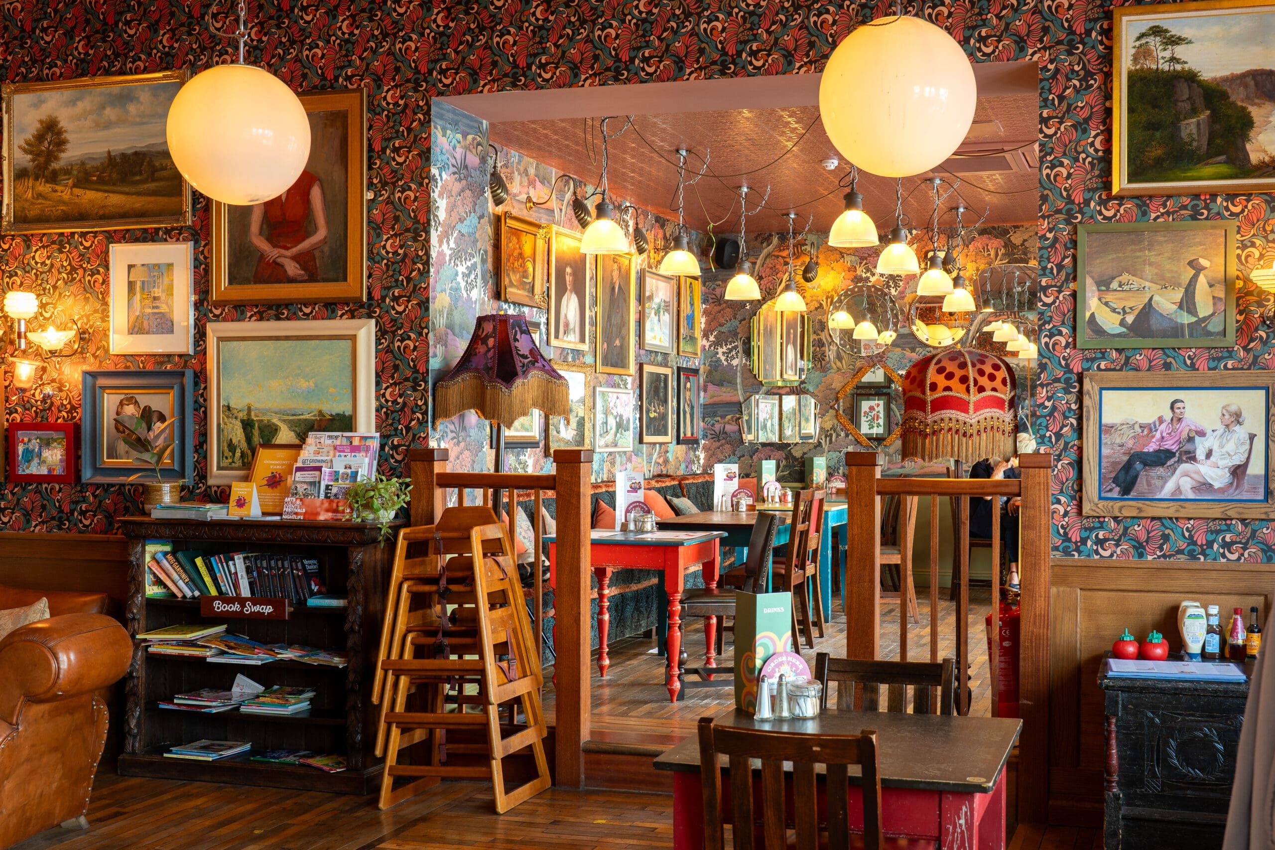 Cozy Porto café with eclectic vintage decor, patterned wallpaper, hanging globe lights, framed art on the walls, wooden tables and chairs, high chairs, and a small bookshelf in the corner.