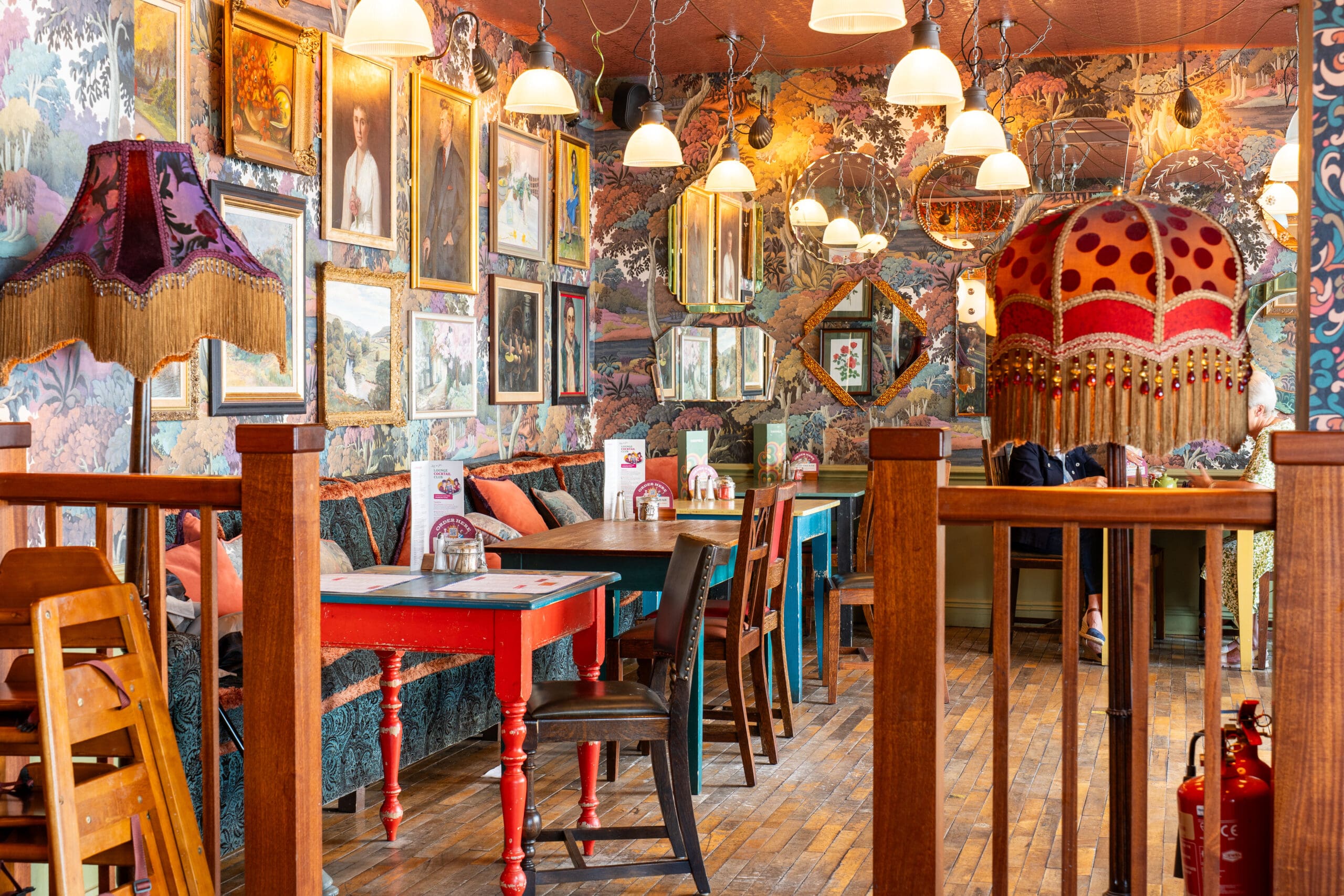 A colorful, eclectic café interior in Porto with vintage furniture, patterned wallpaper, framed art on the walls, mismatched tables and chairs, and unique lampshades creating a cozy, artistic atmosphere.