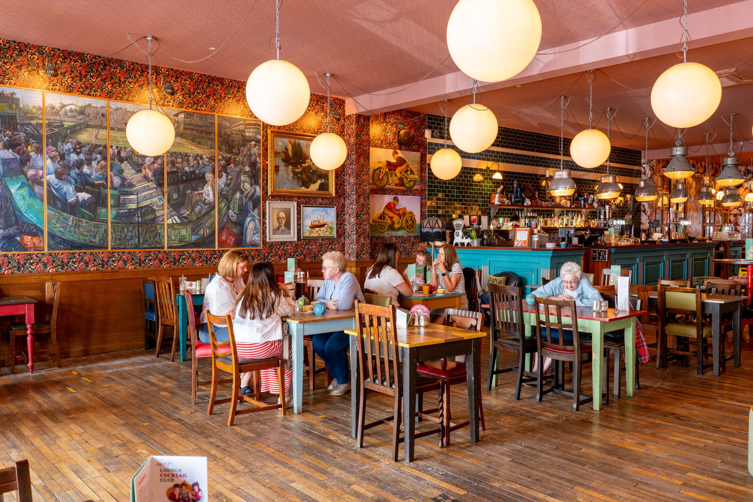 A brightly lit restaurant in Porto with wooden floors, colorful walls, hanging round lights, and eclectic decor. Several groups of people are seated at tables, eating and chatting. The bar area is visible in the background.