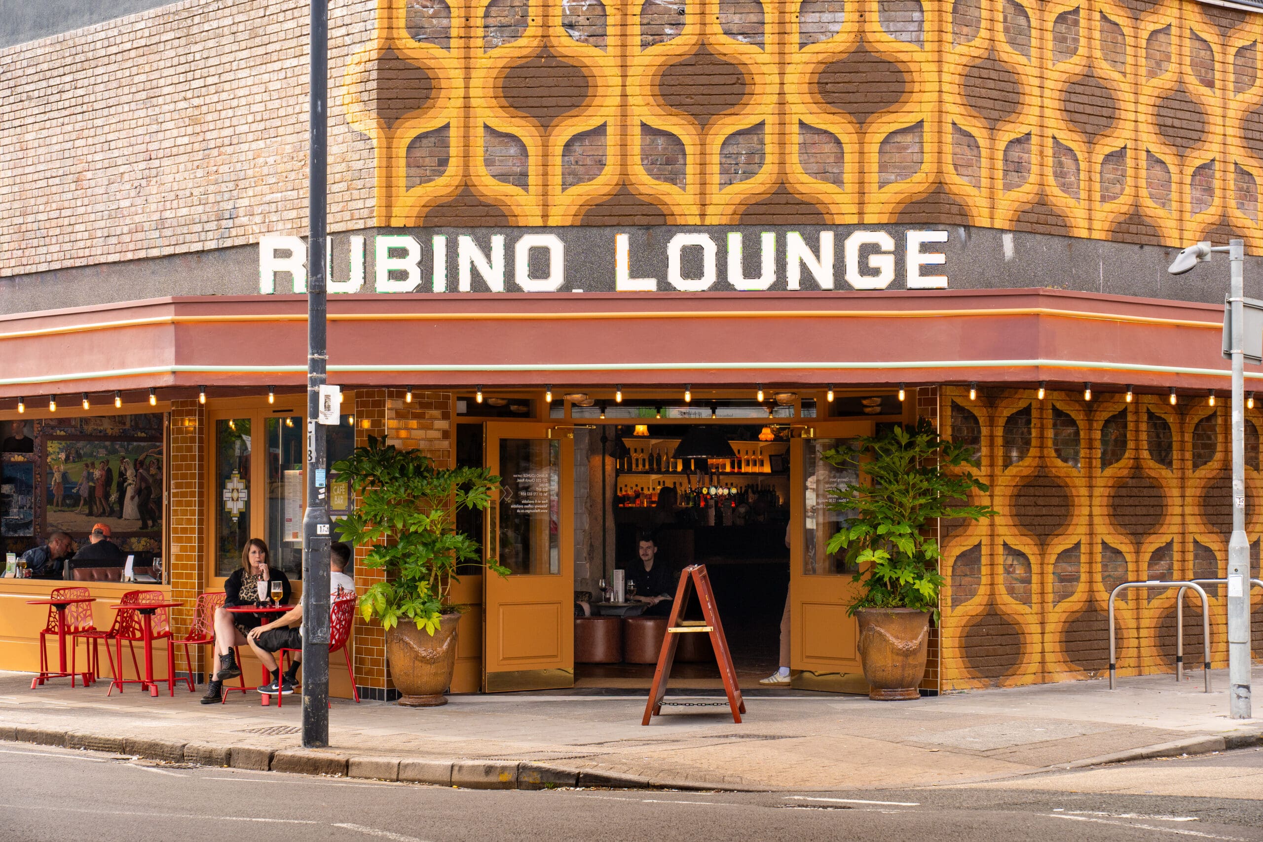 The exterior of Rubino Lounge, featuring retro yellow patterned tiles, large potted plants, red outdoor tables and chairs, and a marinero relaxing outside on a lively city street.
