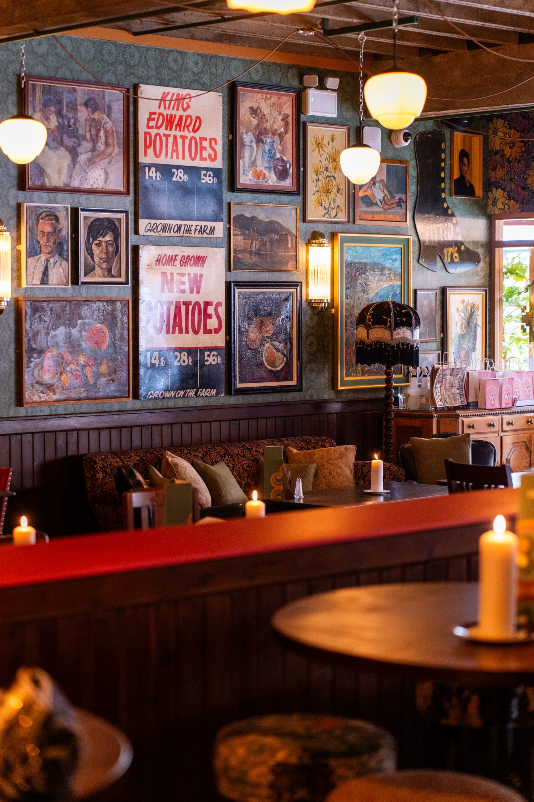 A cozy, dimly lit café with candles on tables, vintage decor, and a gallery wall featuring paintings and retro marinero potato advertisements. Plush cushions and eclectic furniture enhance the warm, inviting atmosphere.