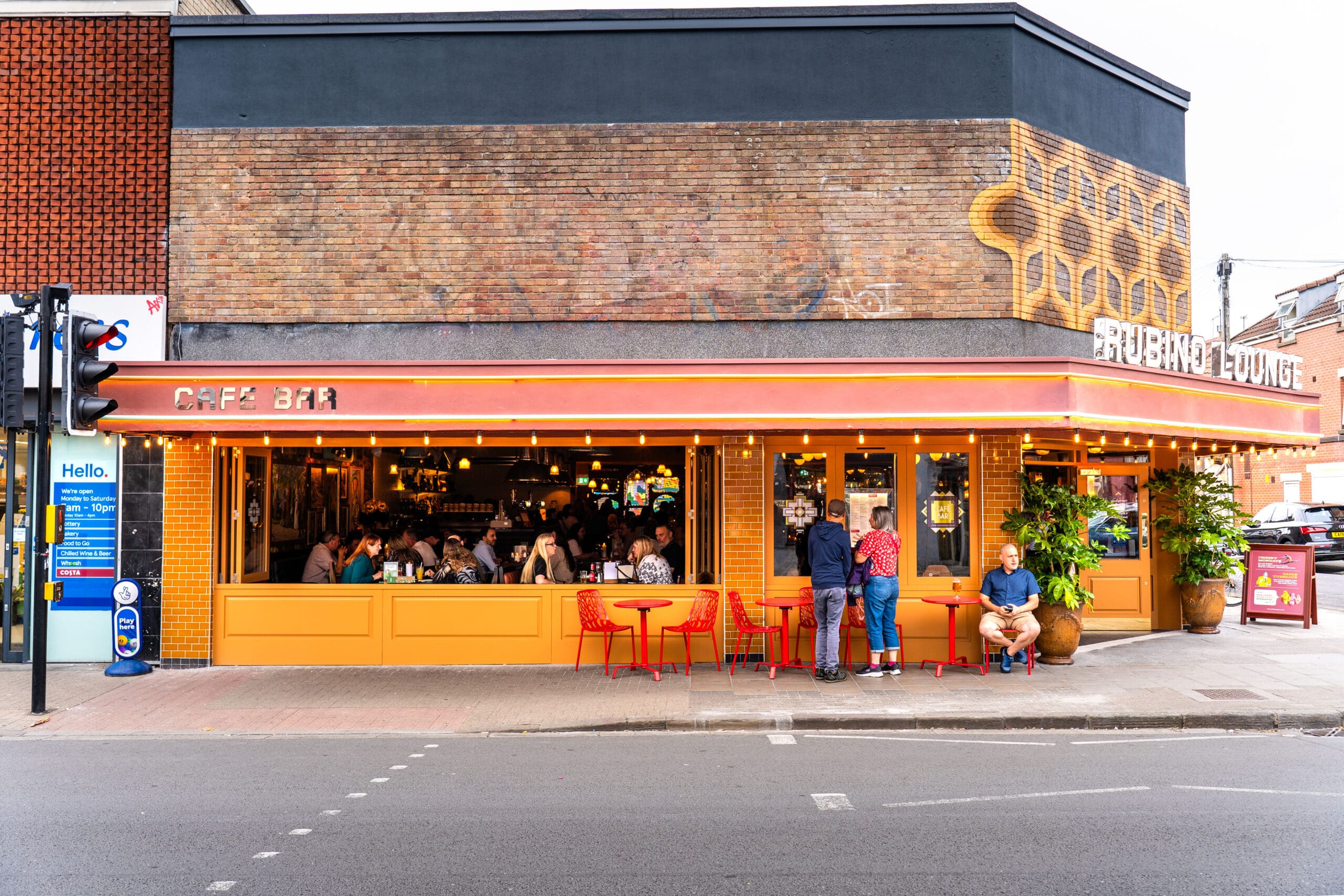 A lively café bar with a marinero vibe, orange and yellow exterior, large windows, and people sitting at red tables outside. More patrons are visible inside under warm lighting. The street and neighboring buildings are also visible.