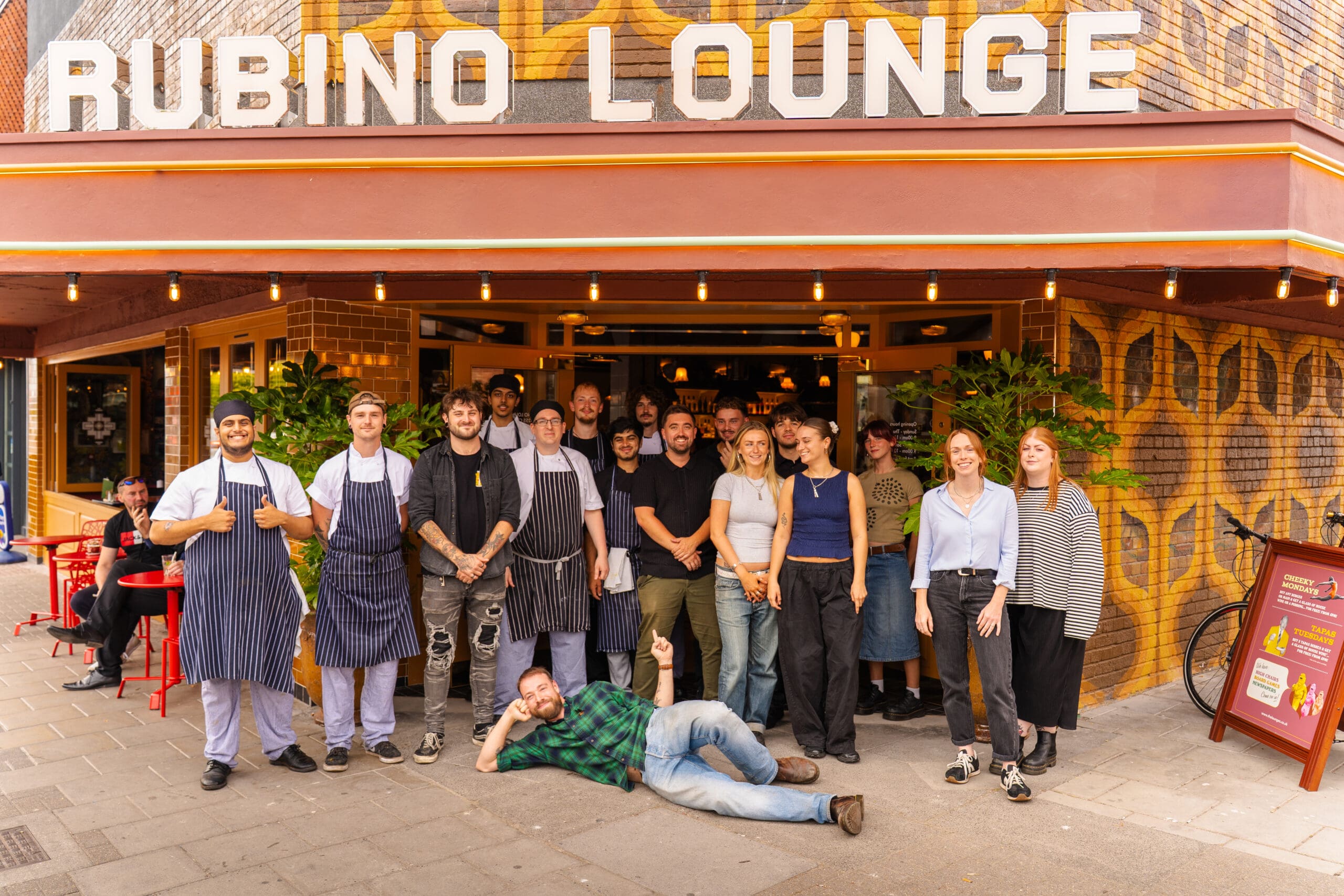 A group of restaurant staff pose and smile outside Rubino Lounge. One person lies on the ground in front, while others stand together under the vibrant Rubino sign. Tables and customers are visible on the bustling sidewalk.