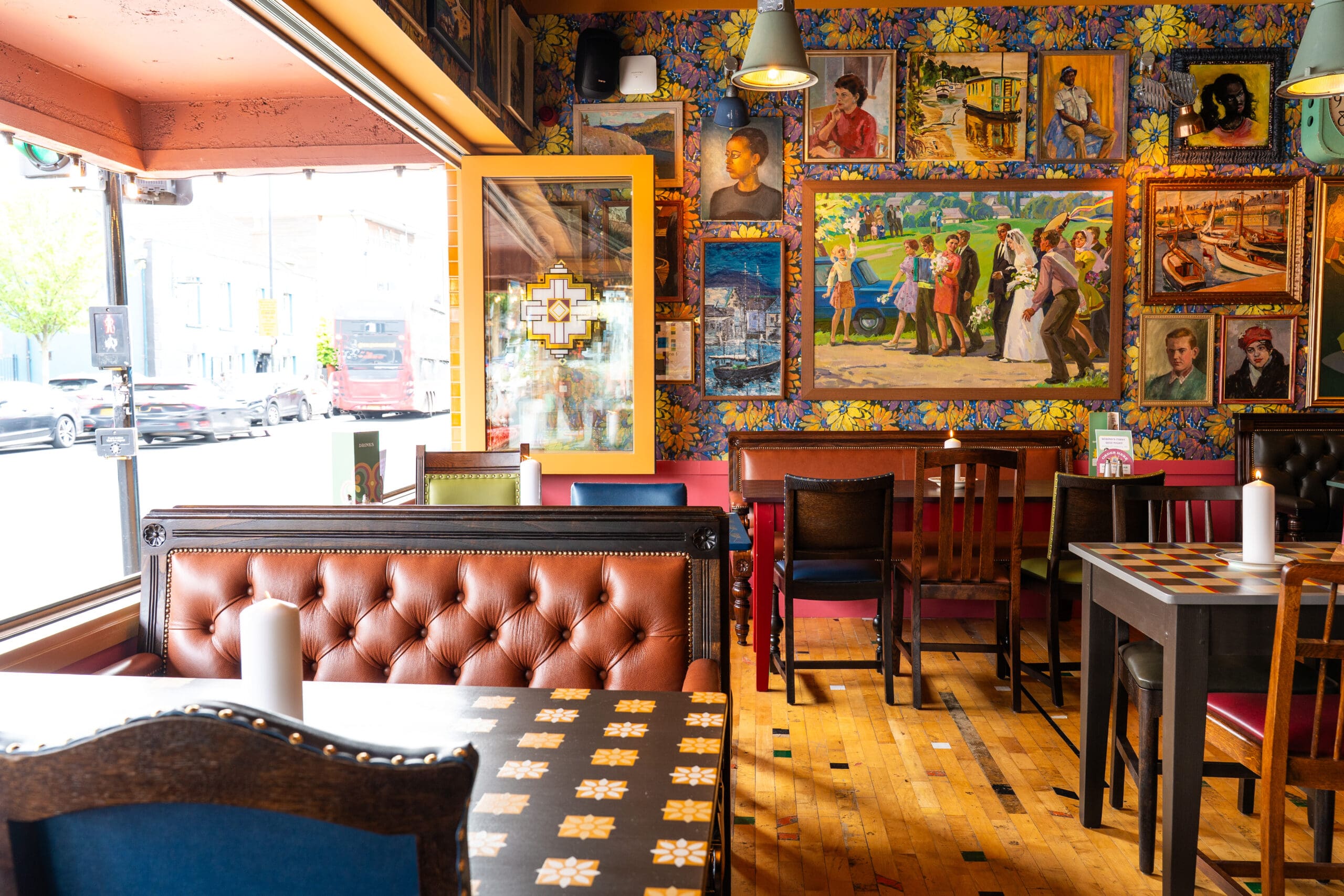 A colorful cafe interior with eclectic furniture, patterned wallpaper, and a wall covered in framed paintings and portraits. Large windows let in natural light, highlighting rubino accents throughout the neatly arranged dining area.
